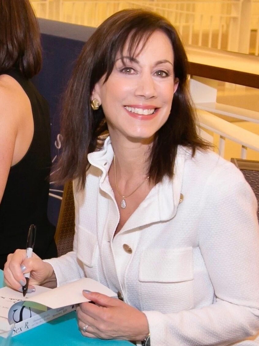 A woman in a white blazer sits at a table, smiling at the camera while signing a book with a pen. Another person is seated beside her, facing away.