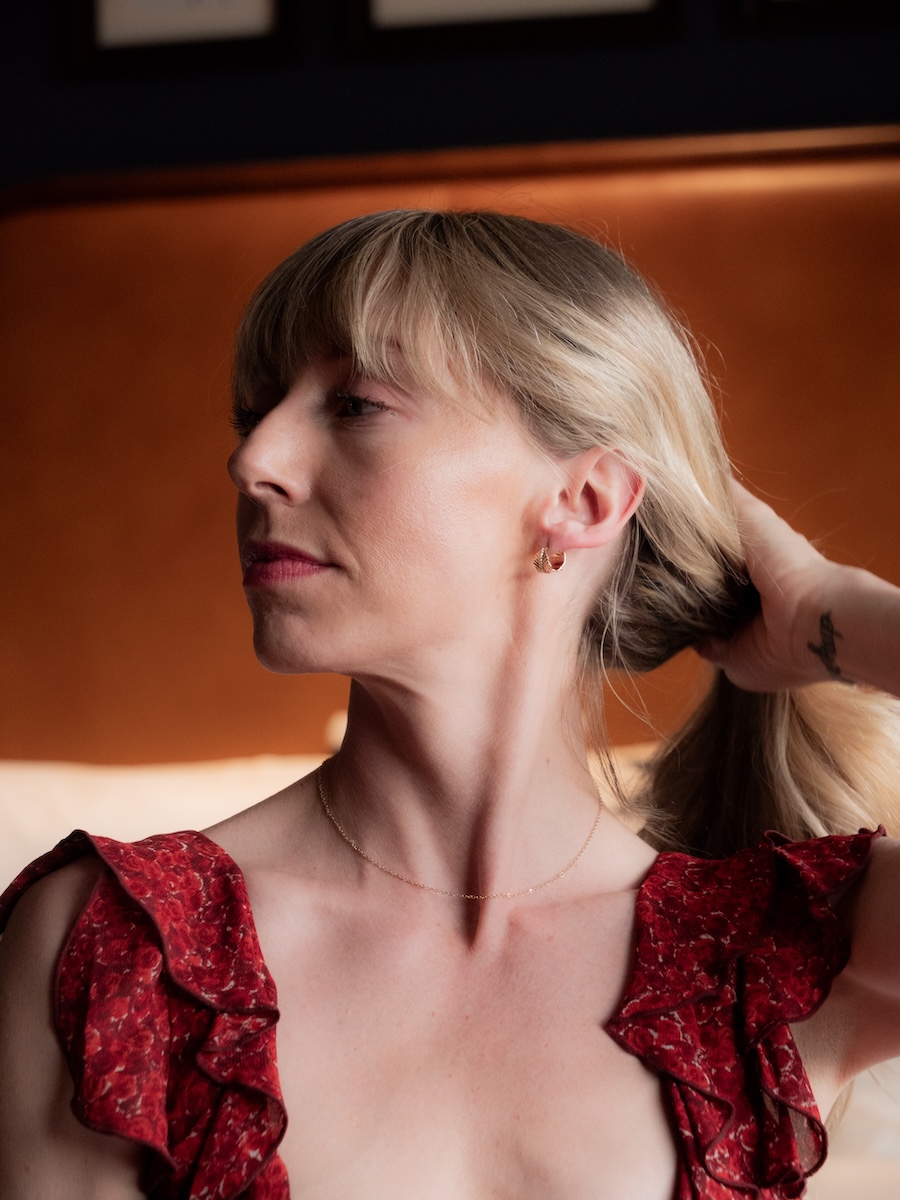 A woman with blonde hair and bangs, wearing a red patterned top with ruffles, looks to the side while holding her hair back. She is in a softly lit indoor setting.