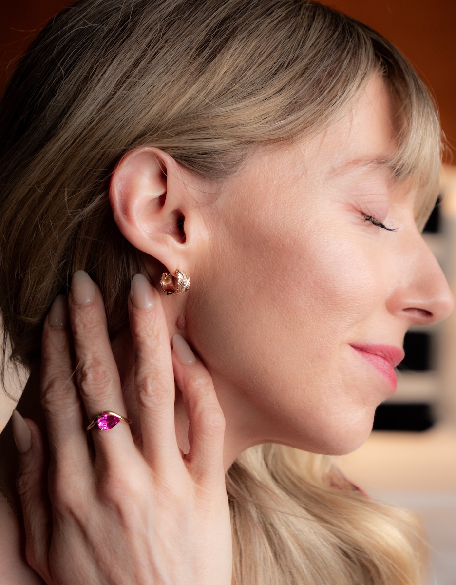 Close-up of a woman with closed eyes showing a gold butterfly earring and a gold ring with a pink gemstone on her hand touching her ear.