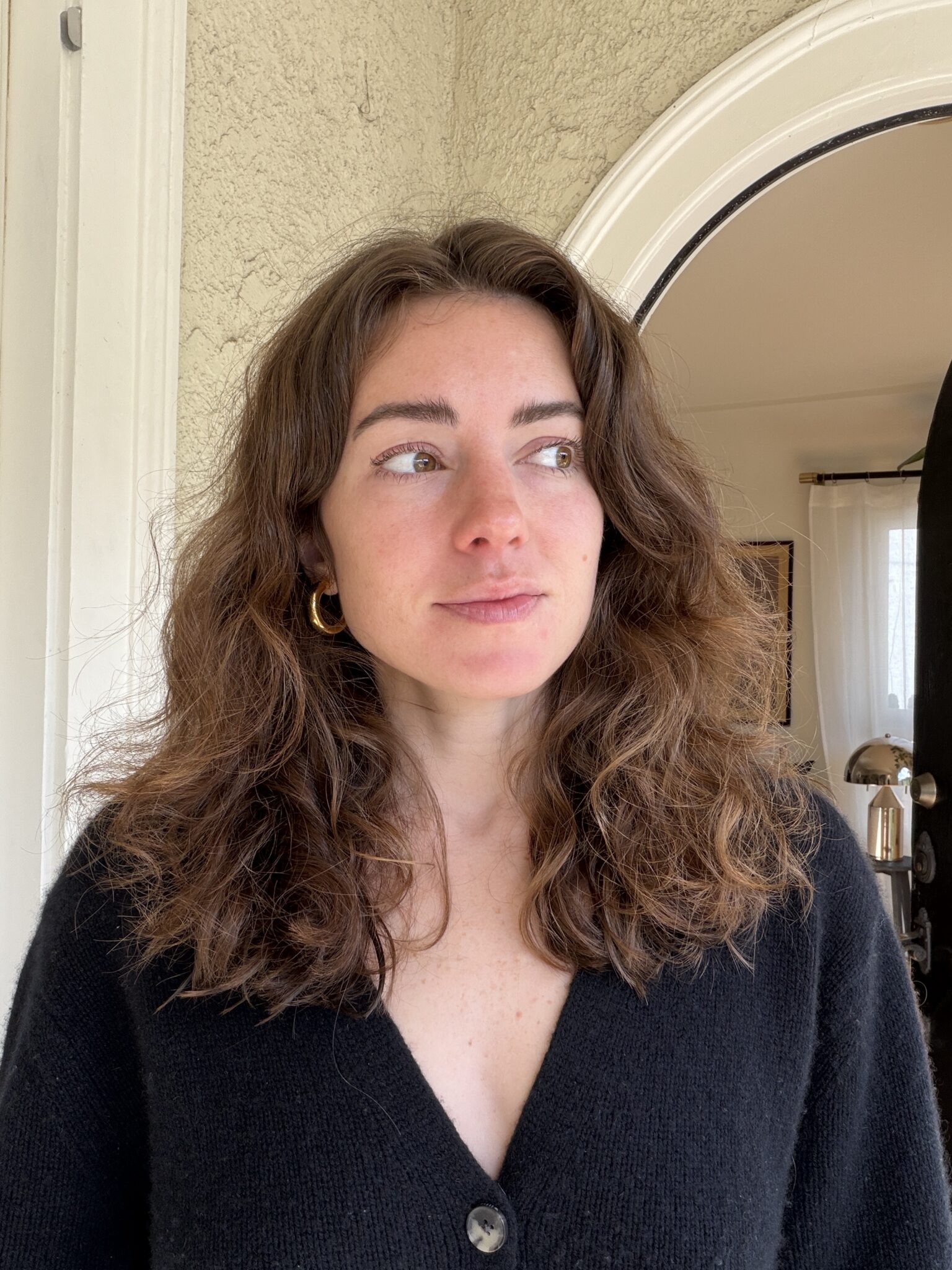 A woman with wavy brown hair wearing a black sweater stands indoors near an arched doorway, looking slightly to the side.