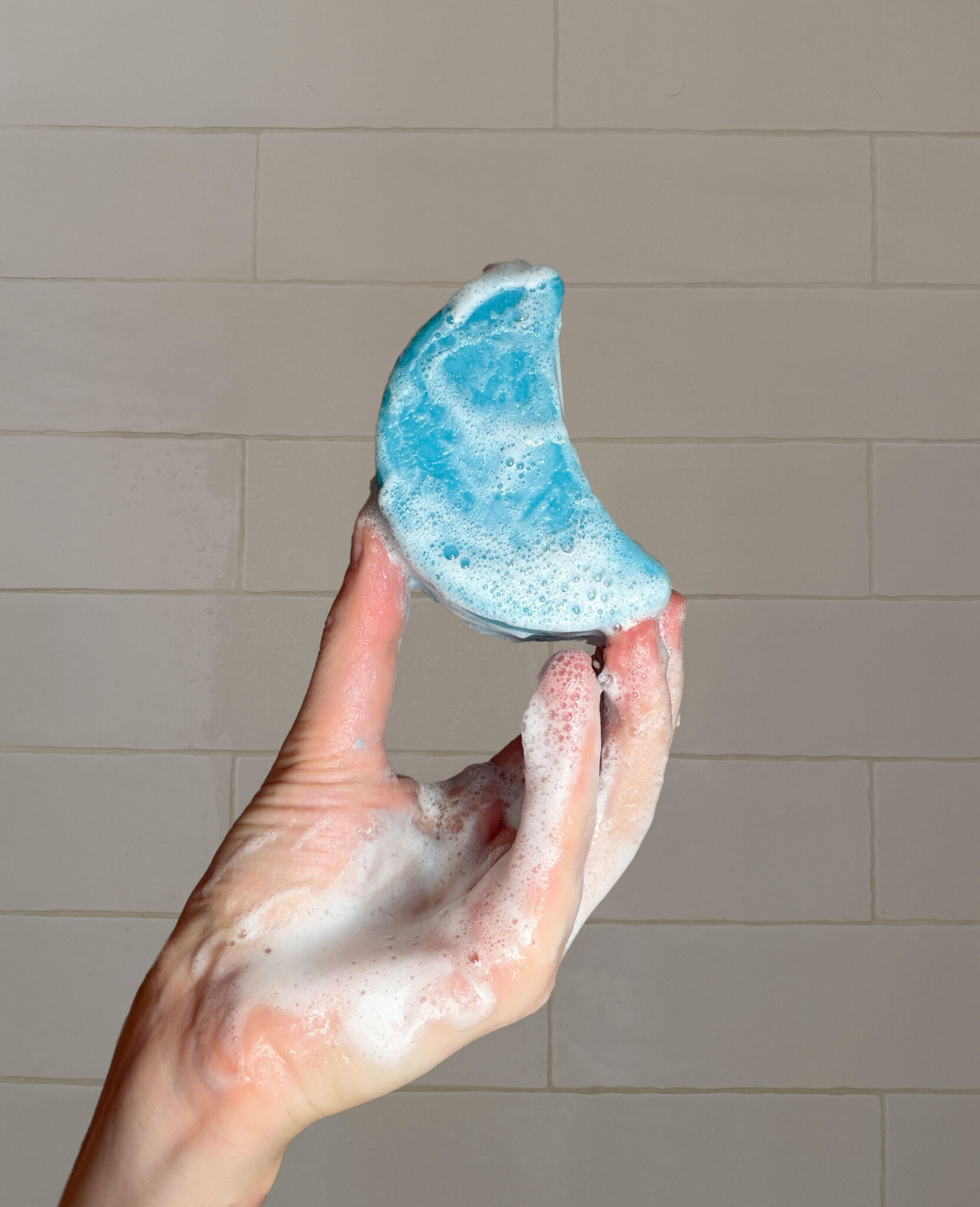 A hand covered in soap suds holds up a blue, crescent-shaped object in front of a tiled wall.