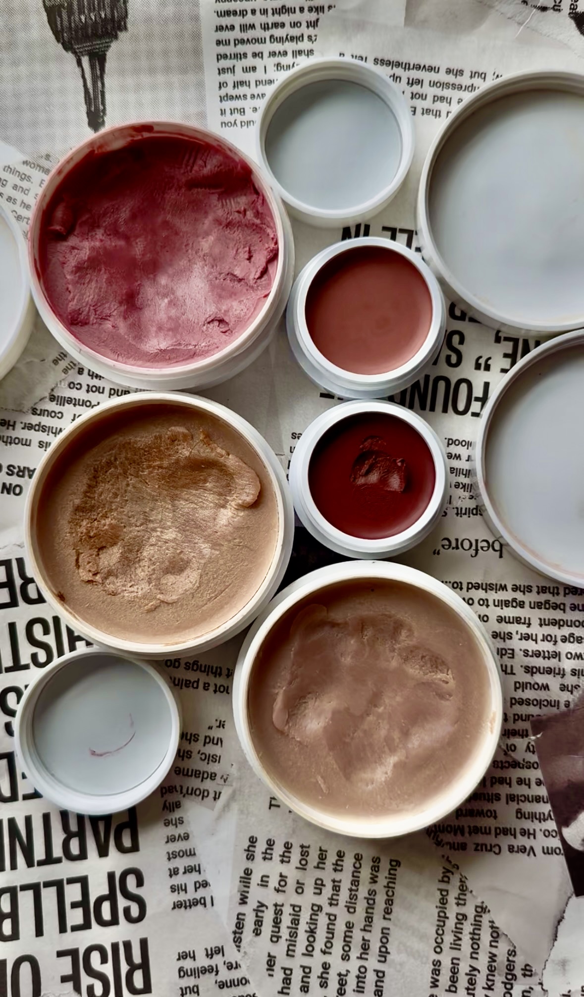 Open containers of various Ilia cream makeup products in shades of red, pink, and beige are placed on newspaper pages with black text.