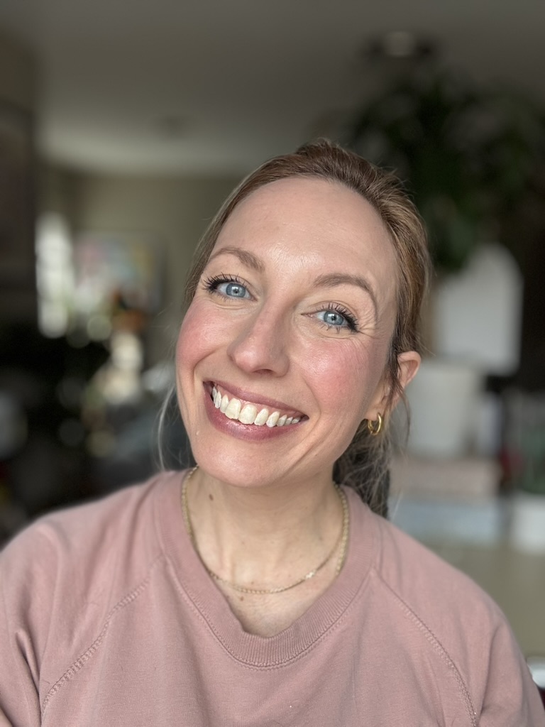 A woman with blue eyes and blonde hair smiles at the camera, wearing a pink Ilia sweatshirt and gold jewelry, indoors with a blurred background.