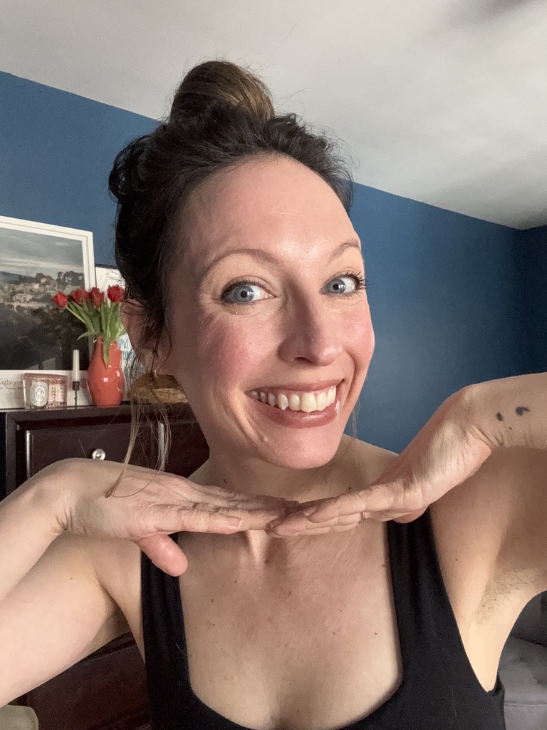 Woman smiling at the camera, holding her hands under her chin in a playful pose, wearing a black Ilia top, with a blue wall and flowers in the background.