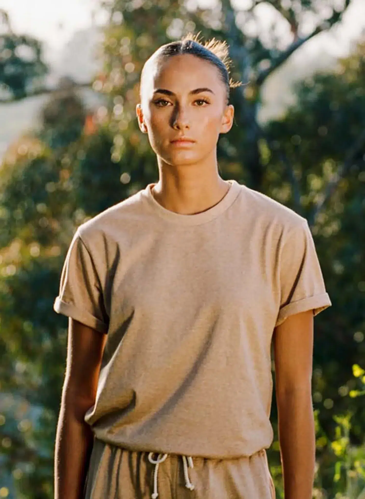 A person with pulled-back hair, wearing a beige t-shirt and drawstring pants, stands outdoors with trees and sunlight in the background.
