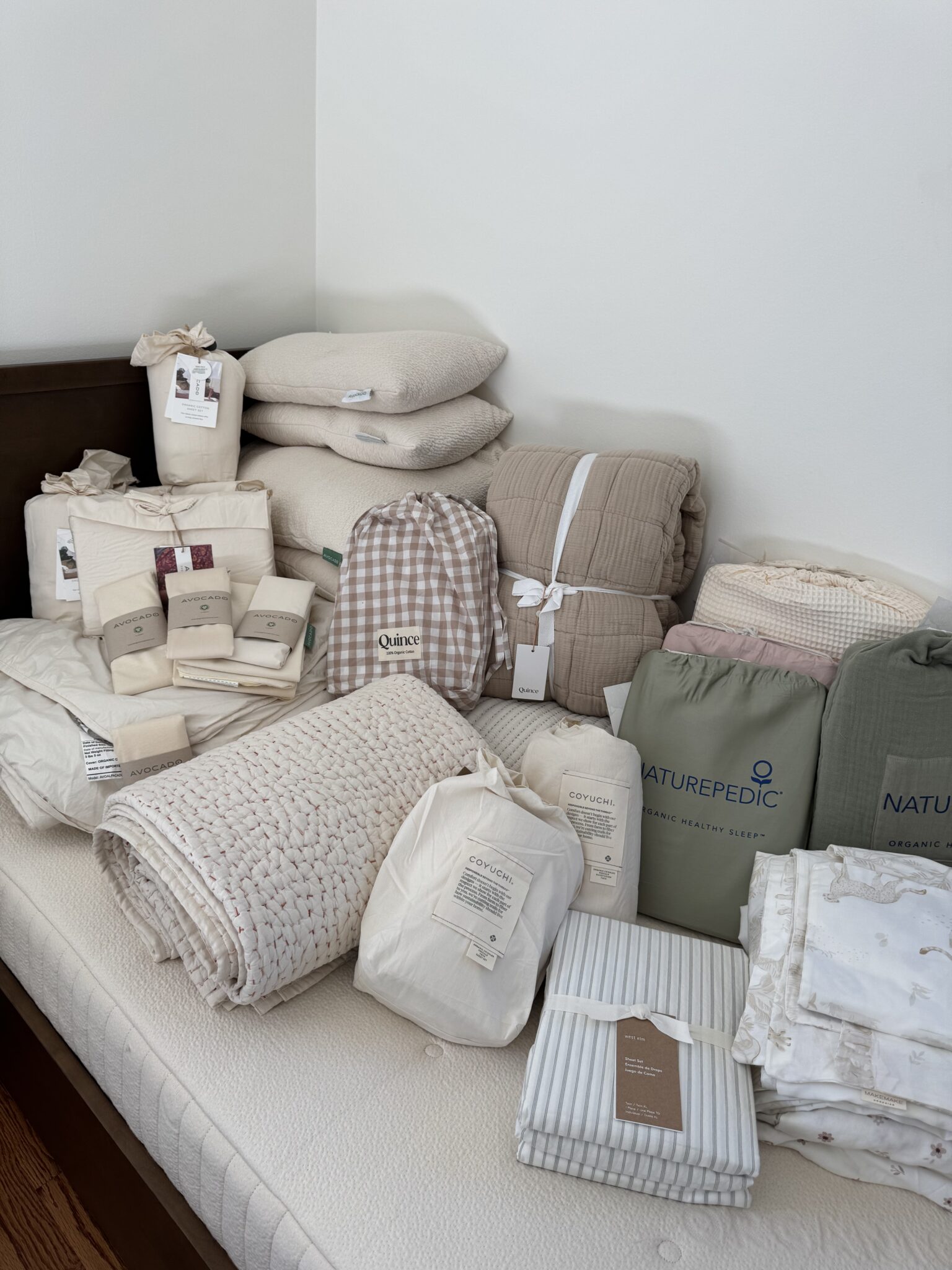 A collection of neatly arranged bedding items, including pillows, comforters, quilts, sheets, and mattress pads, displayed on a bare mattress against a white wall.