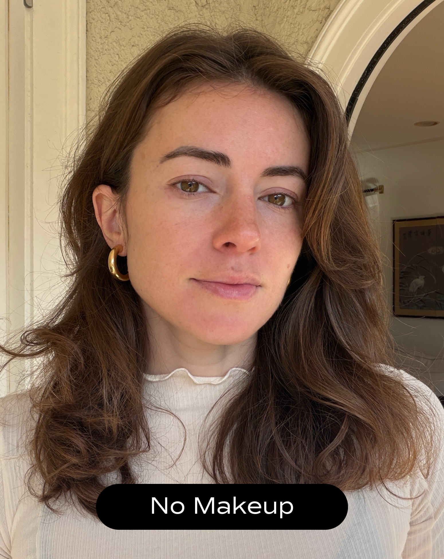 A woman with wavy brown hair and gold hoop earrings stands indoors, wearing a light top. Text at the bottom reads, "No Makeup.