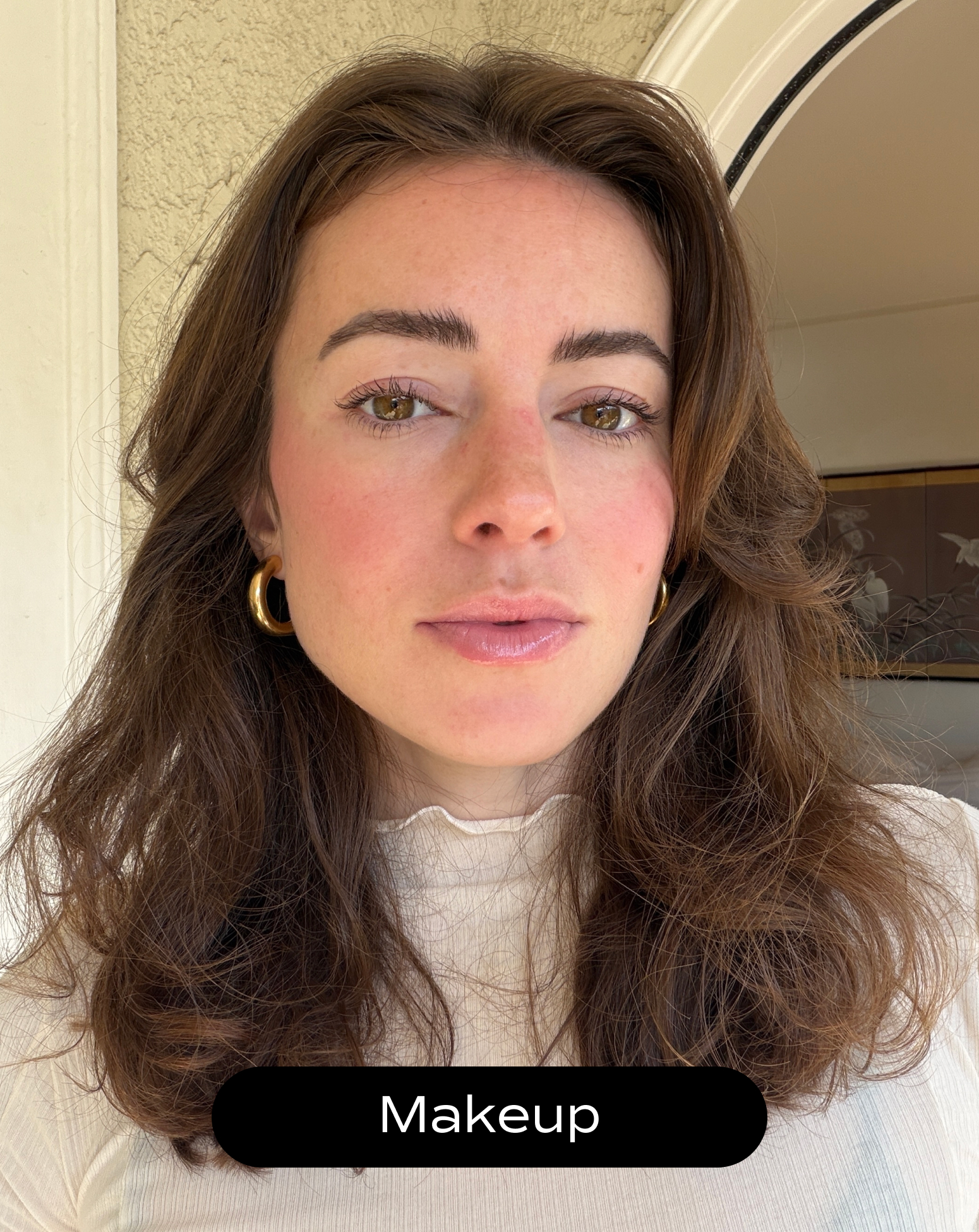 A woman with light skin and wavy brown hair wears natural makeup and gold hoop earrings, standing indoors near a doorway. The word "Makeup" appears at the bottom of the image.