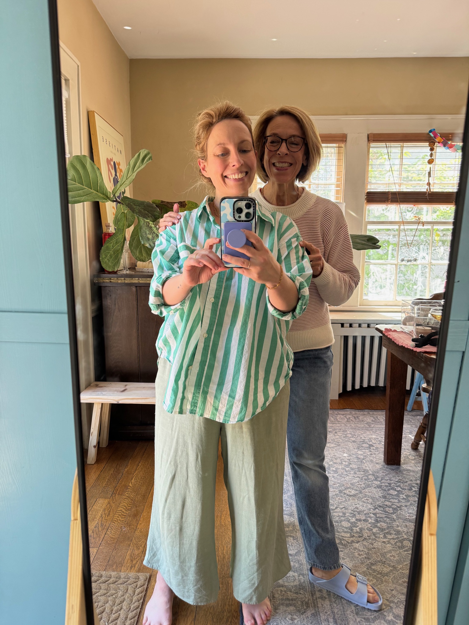Two smiling women stand together in front of a mirror, one taking a selfie with a phone. They are in a sunlit room with plants and framed art visible in the background.