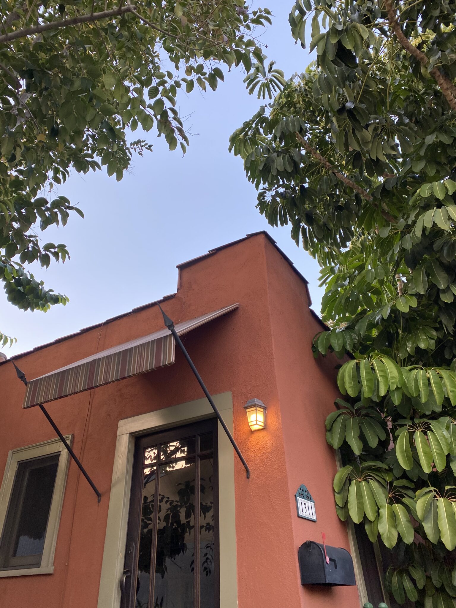 A small orange house with a striped awning, lit porch light, black mailbox, and surrounding green trees under a clear sky.
