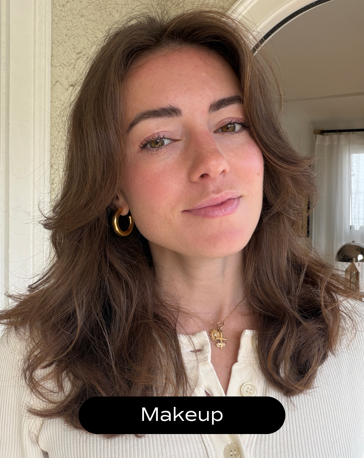 A woman with wavy brown hair and natural makeup is wearing a cream top, hoop earrings, and a necklace, standing indoors near a window.