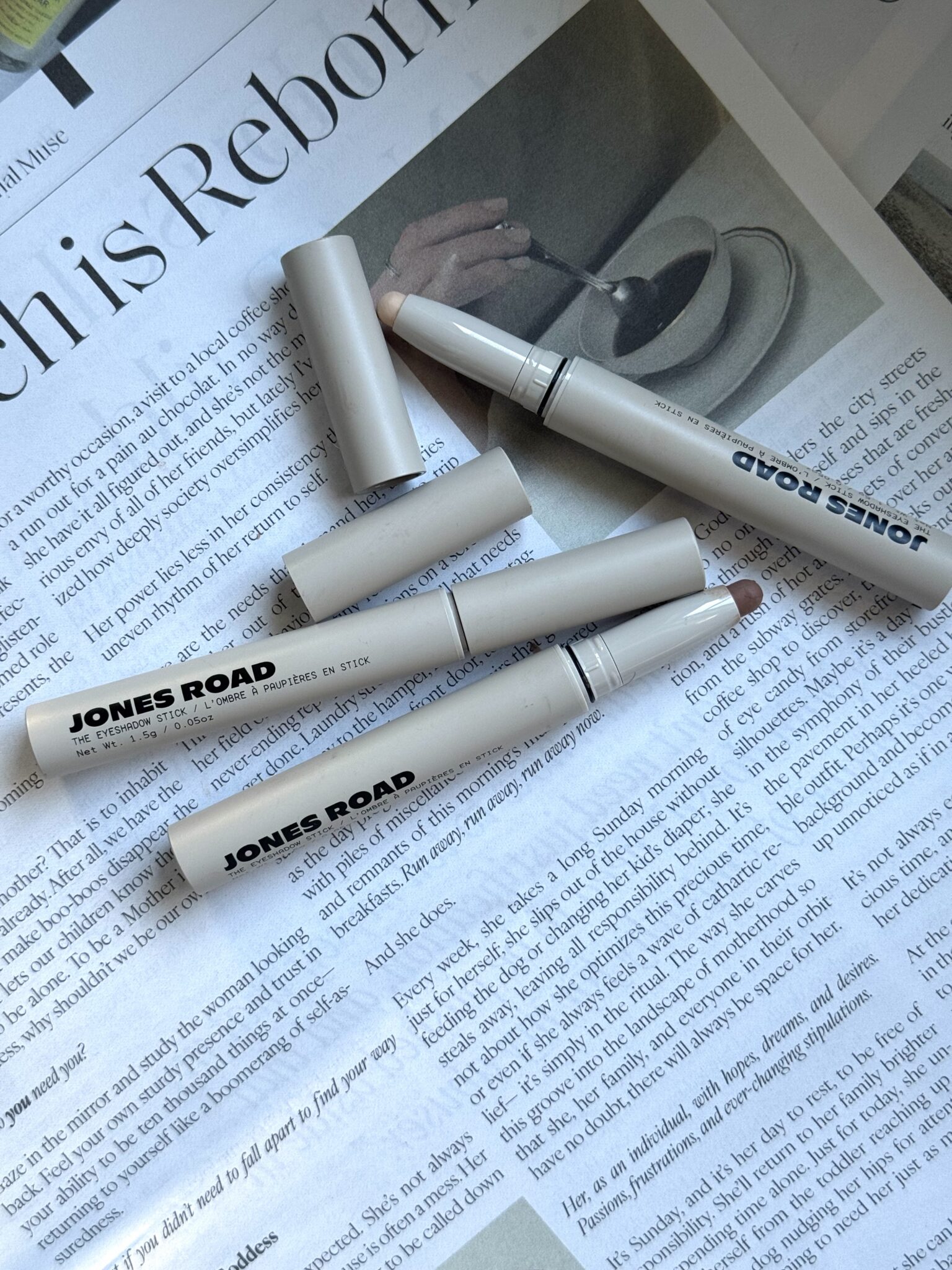 Three Jones Road makeup sticks with caps off are lying on an open newspaper, showcasing the product tips and brand name on the packaging—perfectly complementing your favorite Ilia essentials.