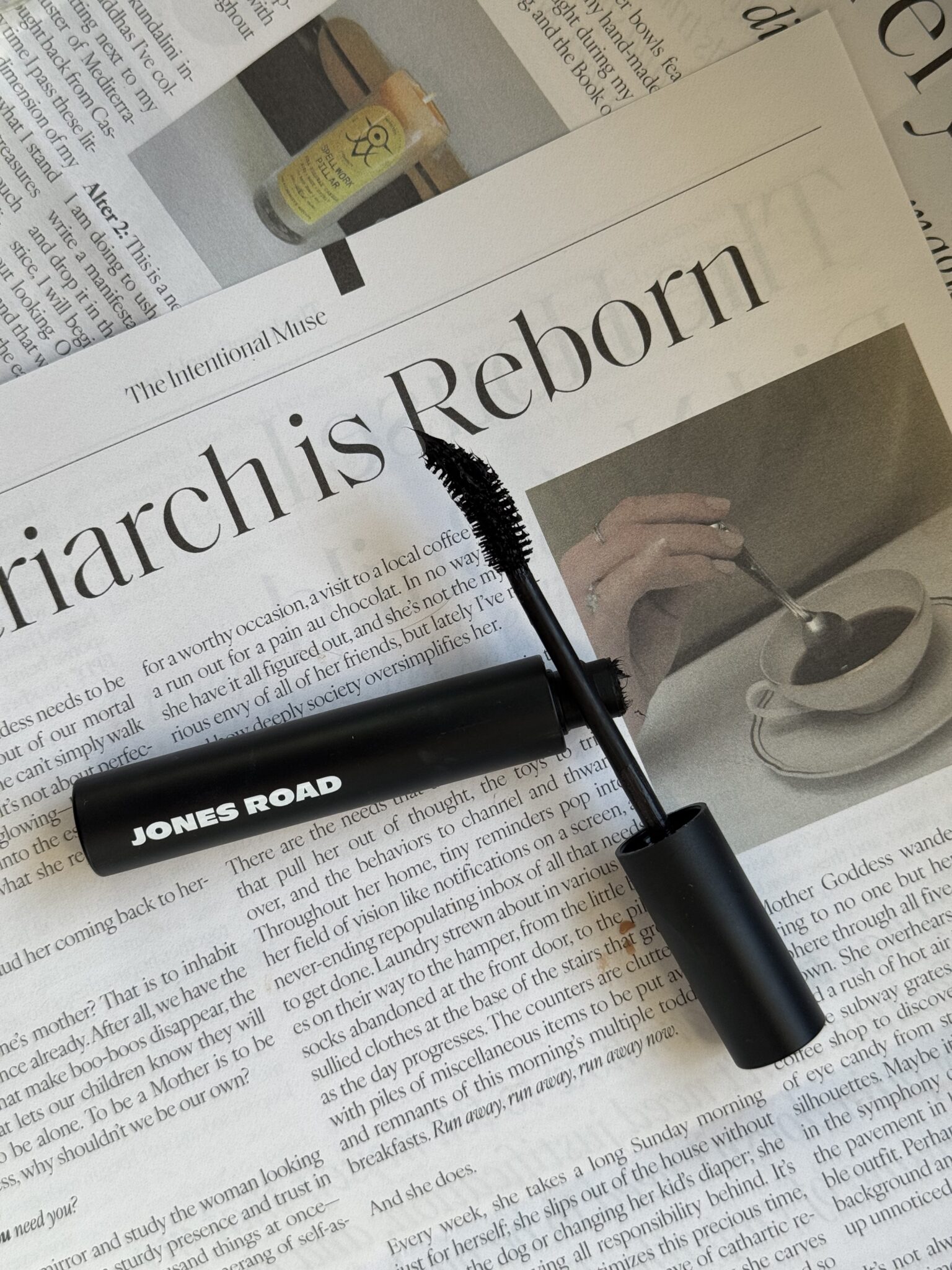 A black JONES ROAD mascara tube with the wand applicator rests on an open magazine alongside Ilia, featuring articles and a photo of a hand holding a cup of coffee.