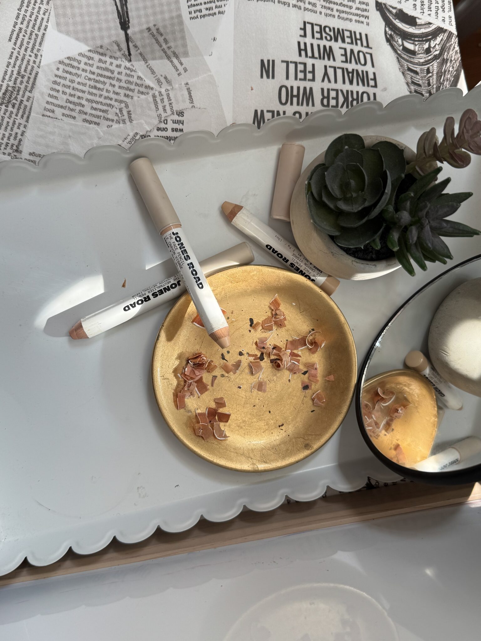 A gold dish with Ilia makeup pencils, pencil shavings, a small mirror, a succulent plant, and newspaper clippings on a white scalloped tray.