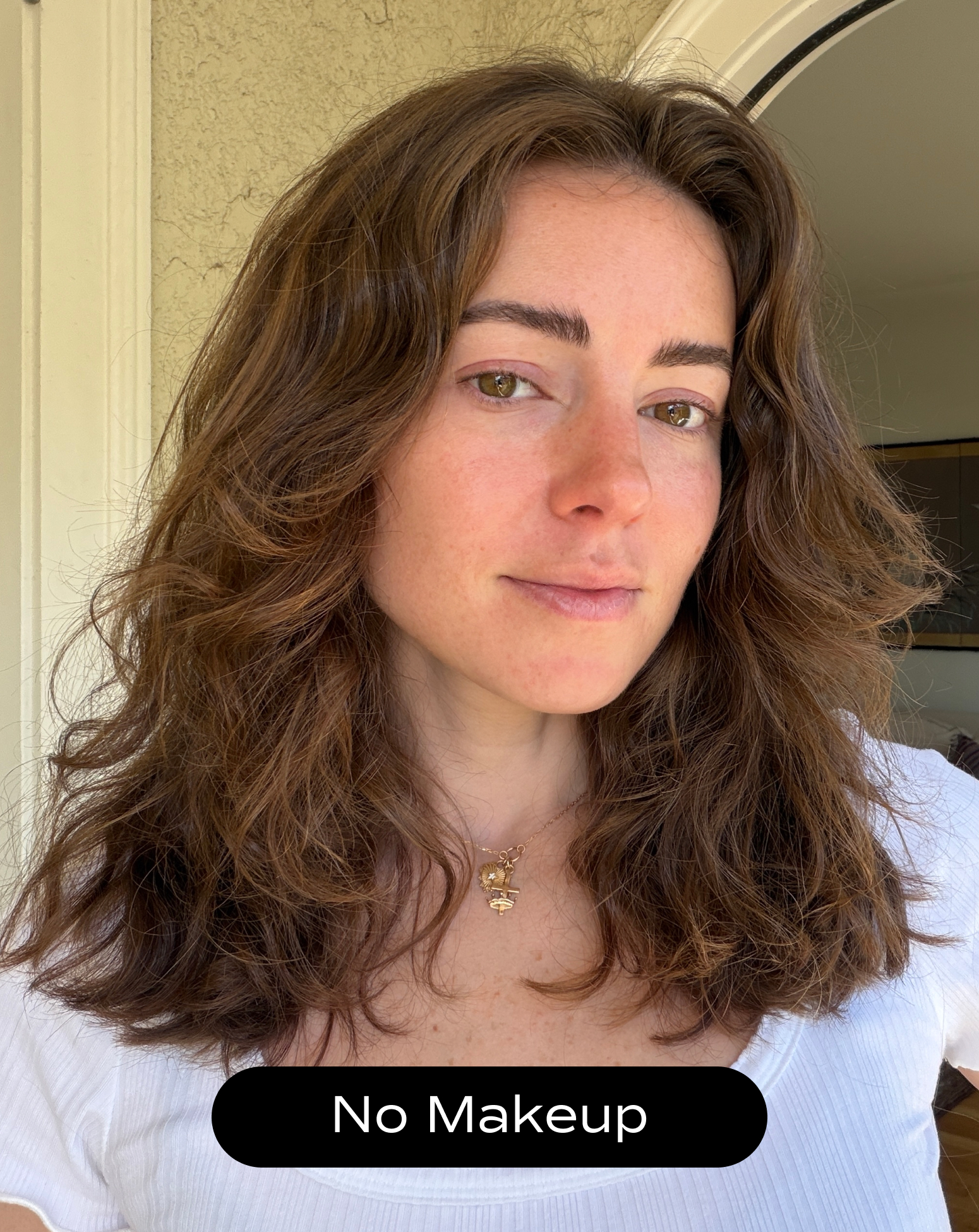 A woman with wavy brown hair and clear skin stands indoors, wearing a white top and a necklace. The text "No Makeup" appears at the bottom of the image.