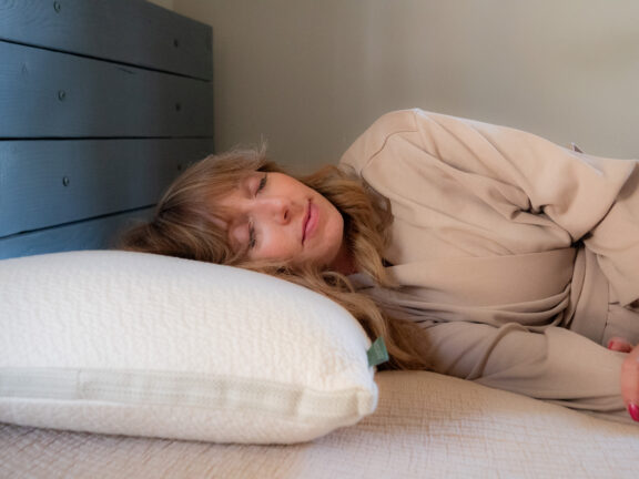 A woman with long hair is lying on her side on a bed, resting her head on a white pillow and wearing a beige robe.
