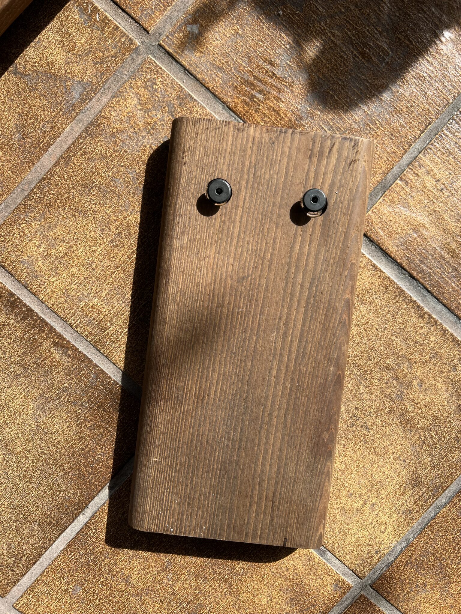 A rectangular wooden plank with two black round screws on top, placed on a brown tiled floor in sunlight.