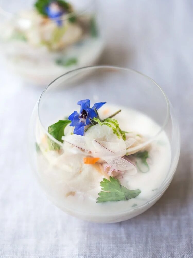 A glass bowl with a creamy white soup garnished with herbs, thinly sliced vegetables, and a blue edible flower on top.