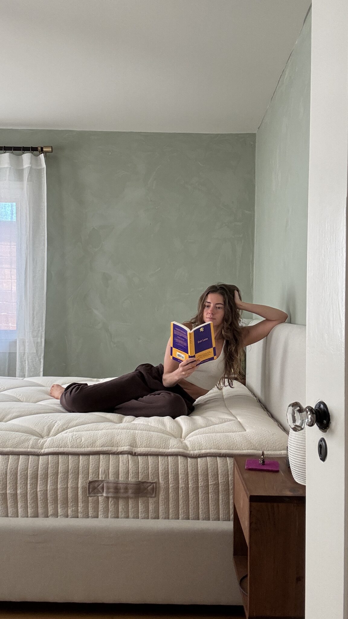 A woman lies on a bed reading a book in a softly lit bedroom with green walls and a window with sheer curtains.
