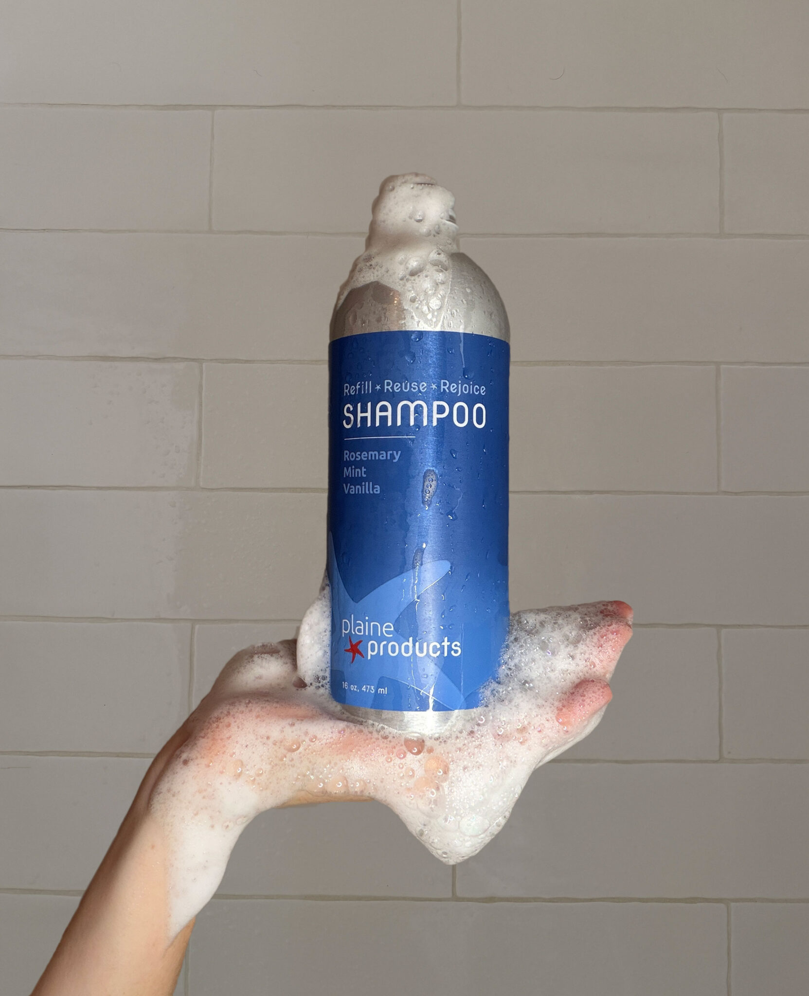 A hand covered in foam holds a Plaine Products shampoo bottle with a blue label in front of a tiled wall.