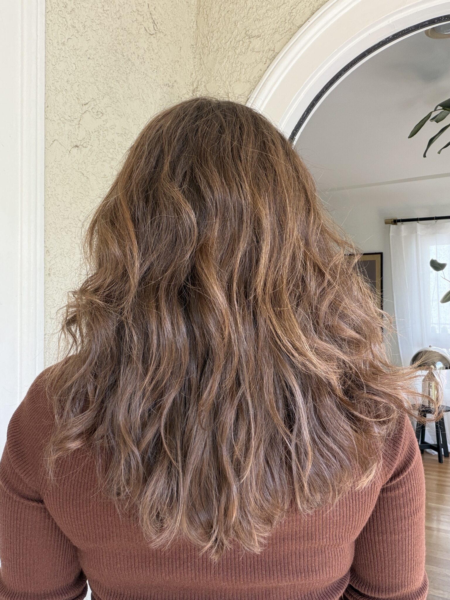 Person with shoulder-length, wavy brown hair wearing a ribbed brown top, standing indoors facing away from the camera.
