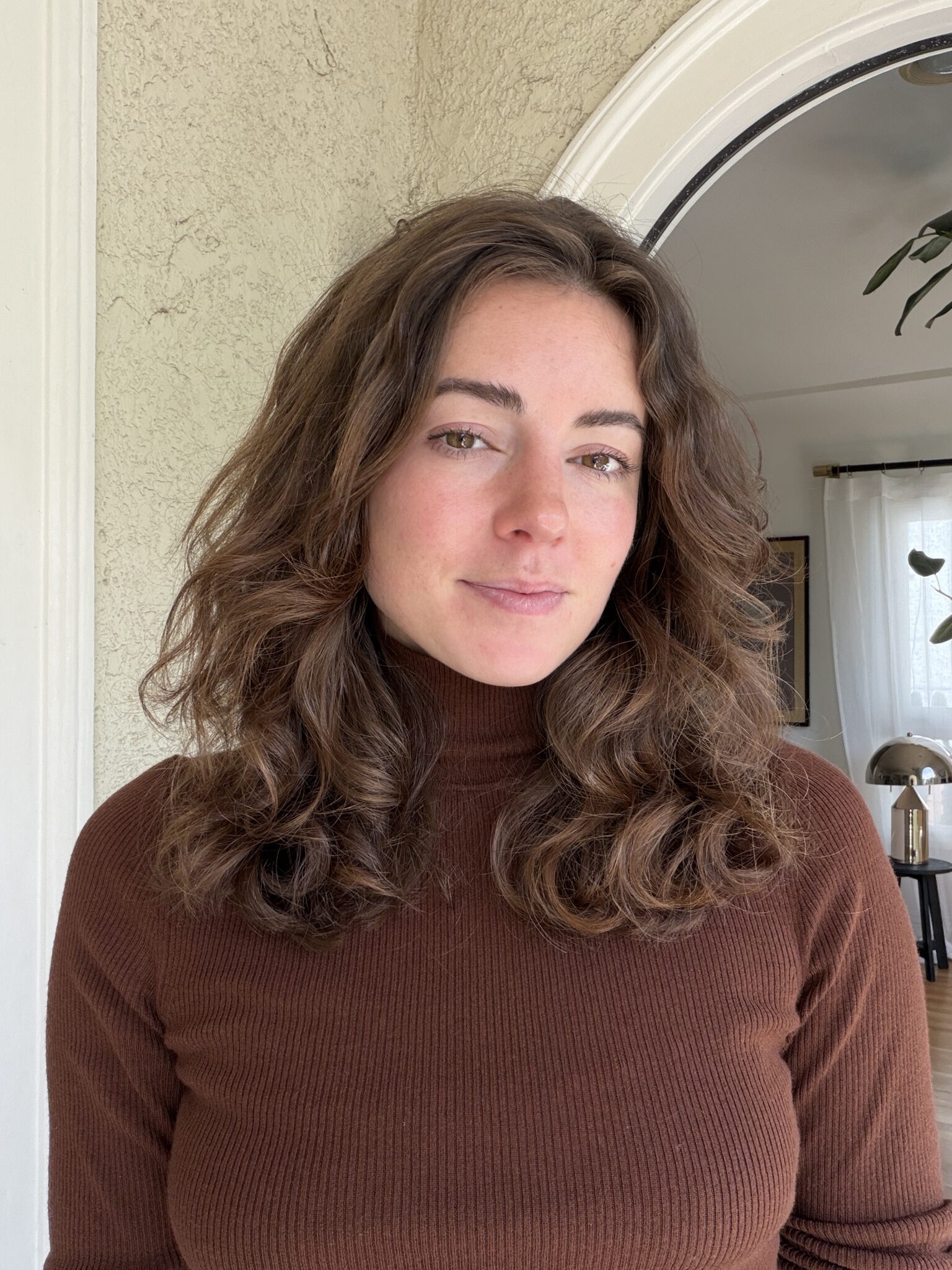 A person with wavy brown hair wearing a brown turtleneck stands indoors in front of a textured wall and arched doorway.