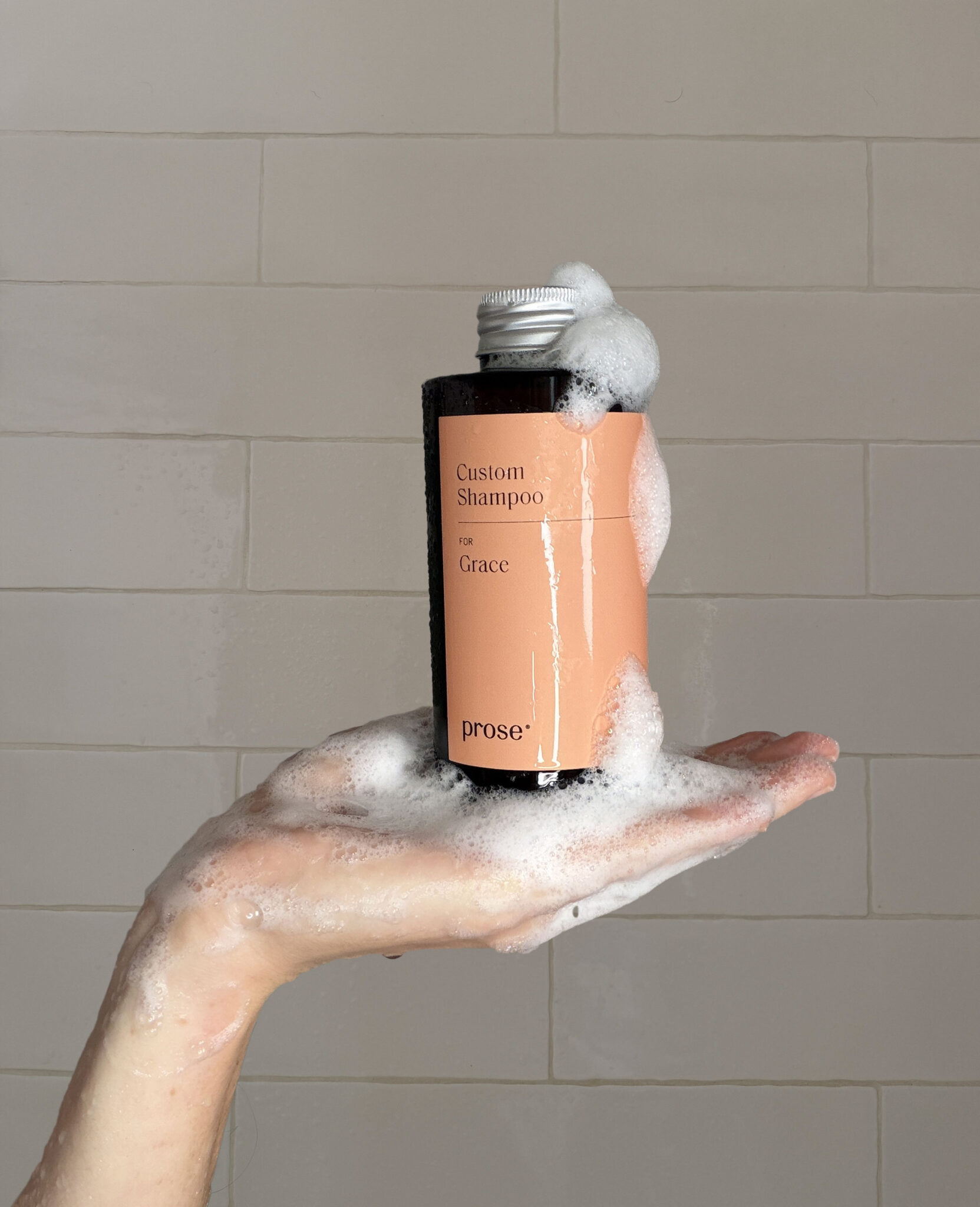 A hand covered in soap holds a bottle of custom shampoo labeled "for Grace" with foamy suds on the cap, against a tiled background.