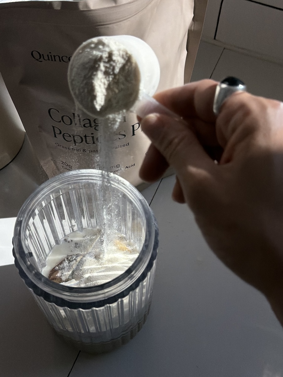 A hand holding a scoop of collagen peptides powder being poured into a blender cup with other ingredients inside.