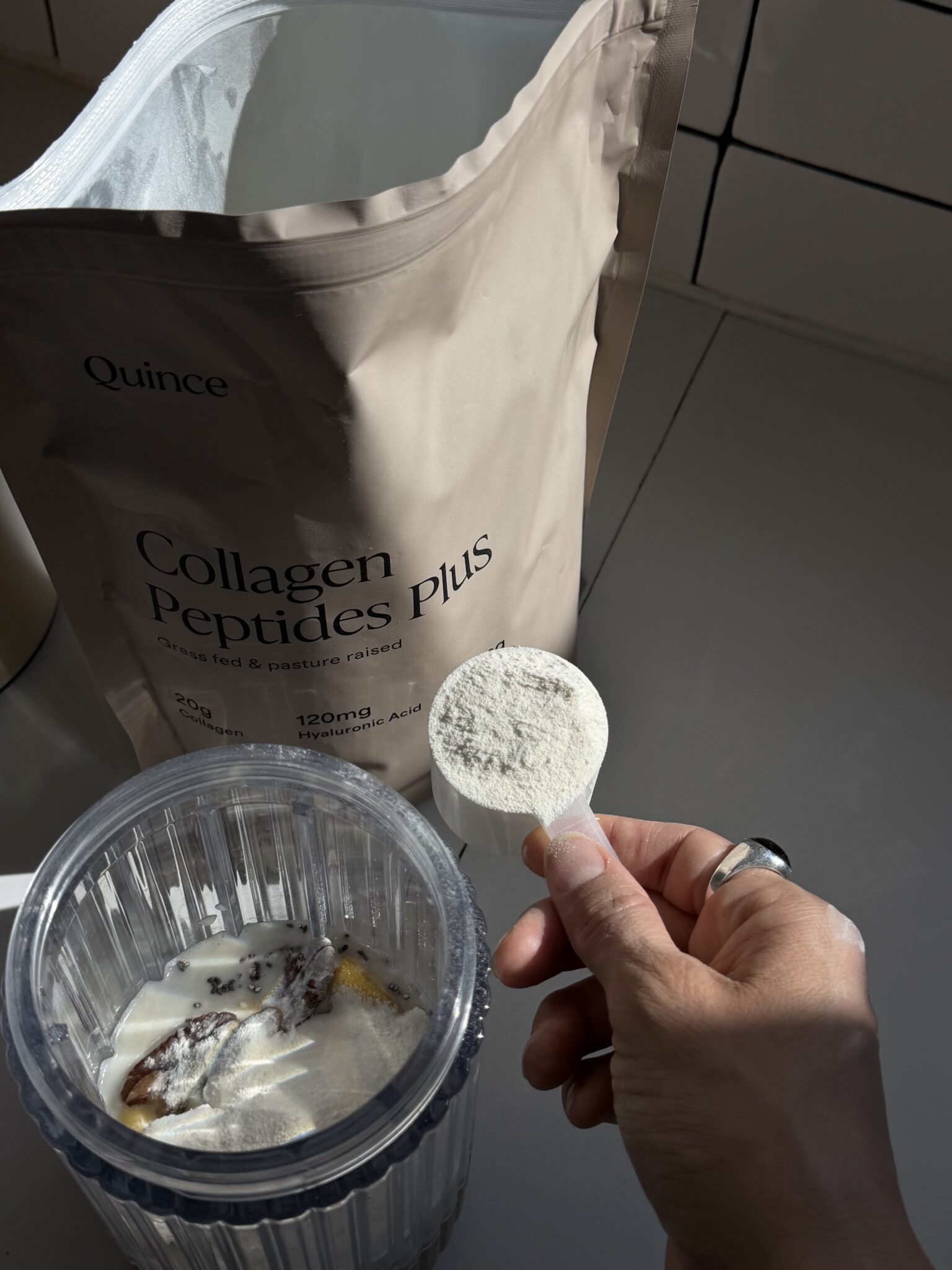 A hand holds a scoop of collagen peptide powder above a blender cup containing milk and banana, with a bag labeled "Collagen Peptides Plus" in the background.