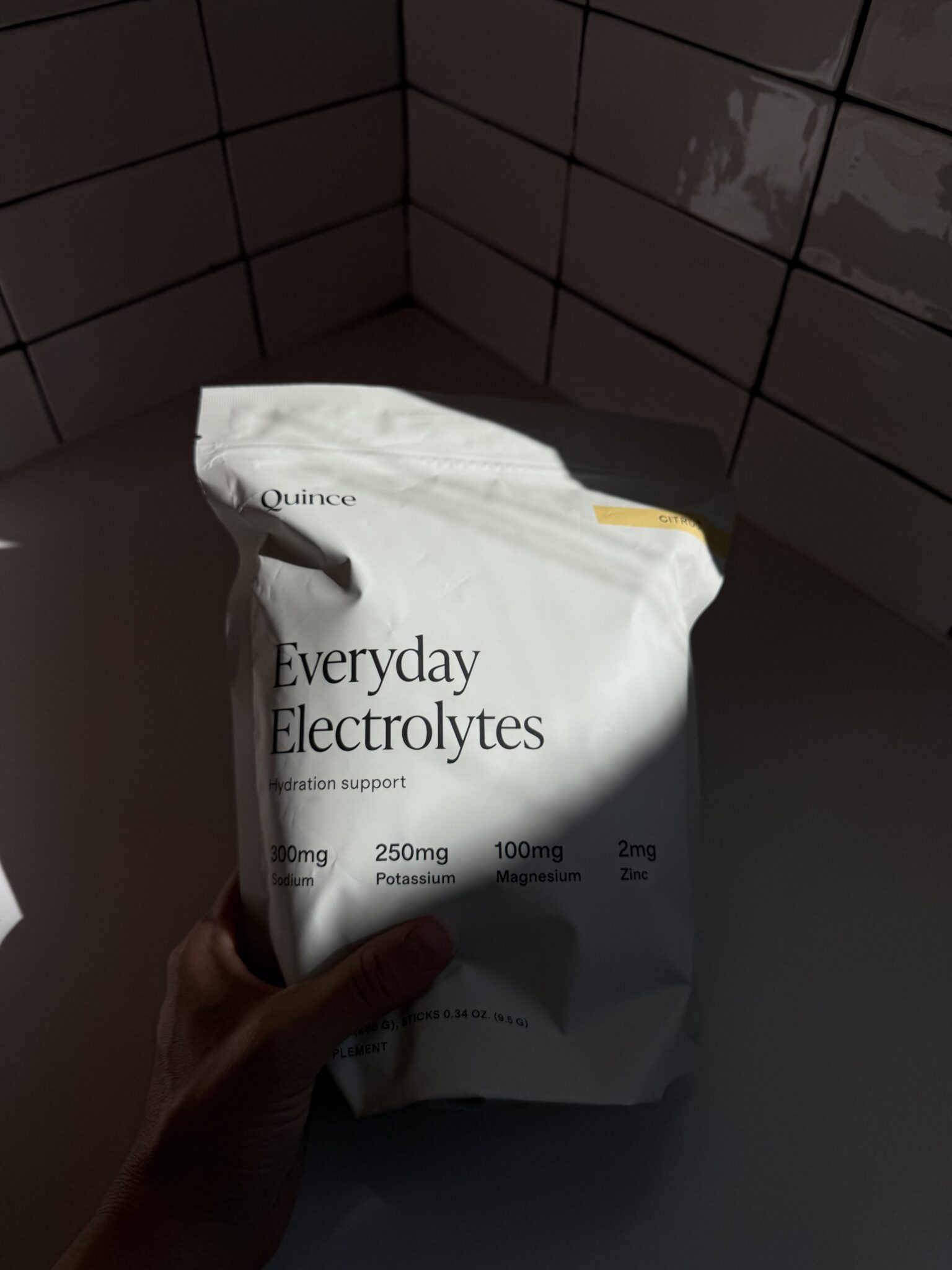 A hand holds a white bag labeled "Everyday Electrolytes" by Quince, listing sodium, potassium, magnesium, and zinc content, against a tiled wall background.