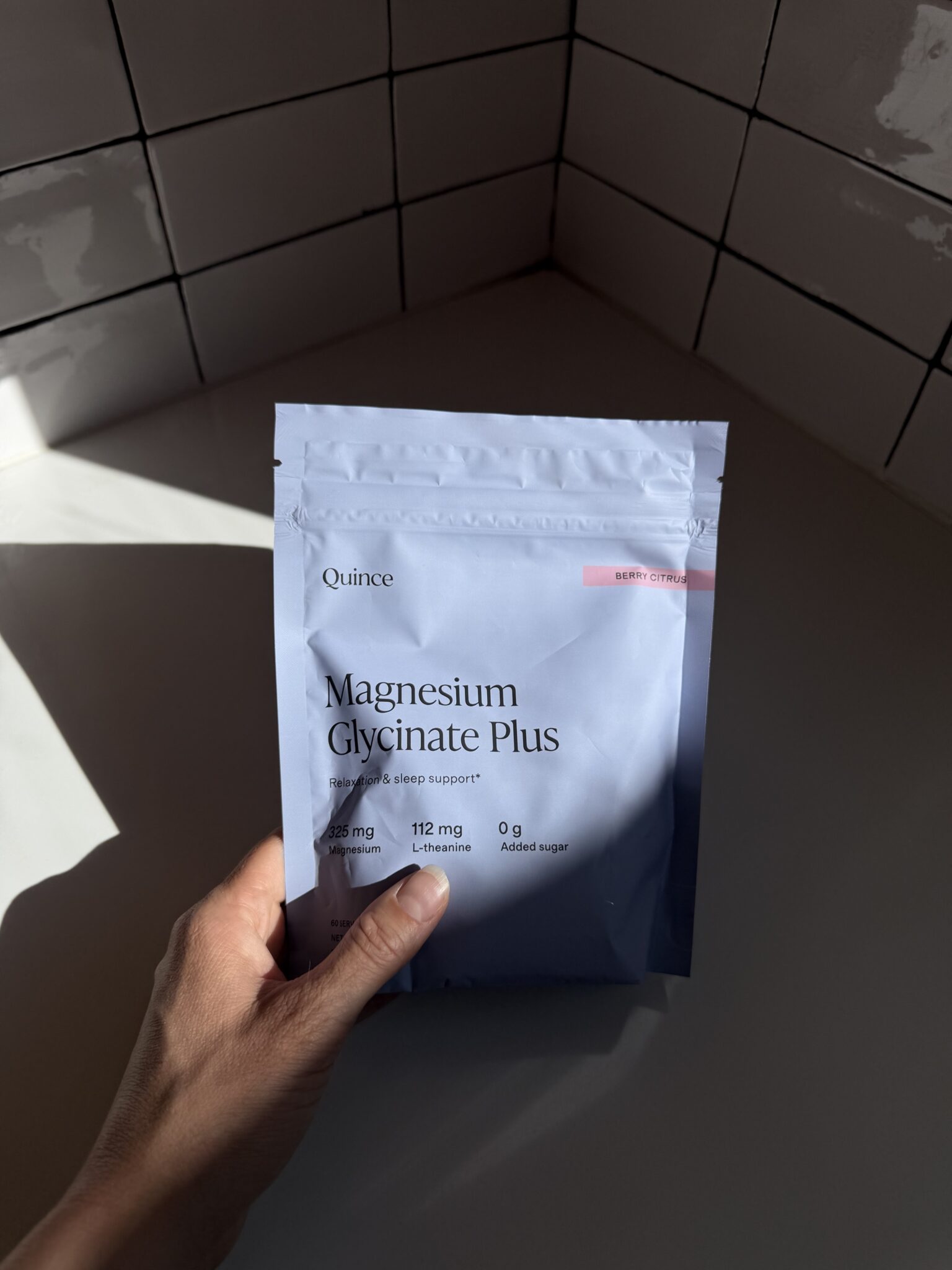 A hand holds a resealable pouch labeled "Magnesium Glycinate Plus" by Quince in a sunlit tiled corner.