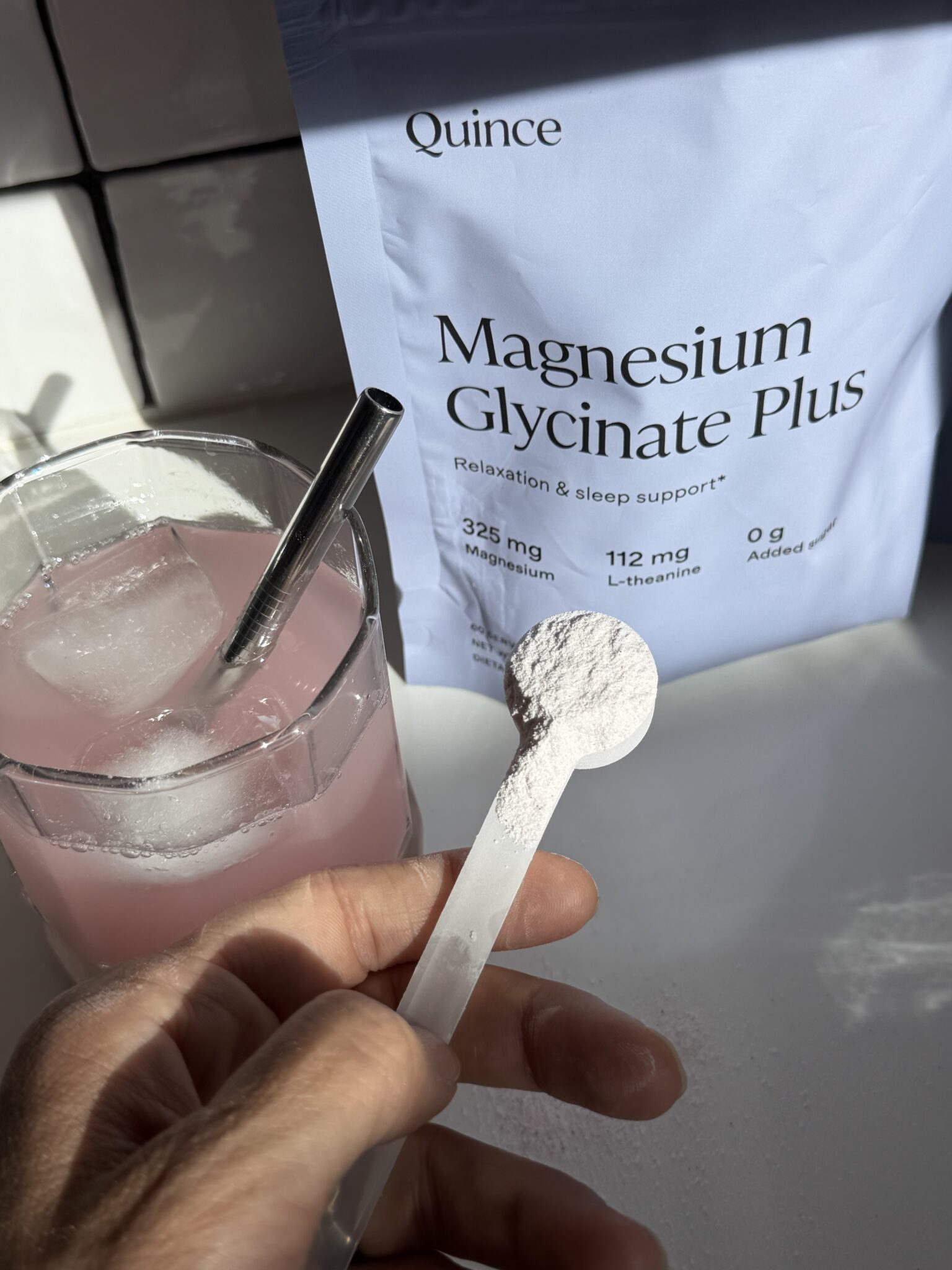 A hand holds a scoop of white powder above a glass of pink drink with ice; a bag labeled "Magnesium Glycinate Plus" is in the background.