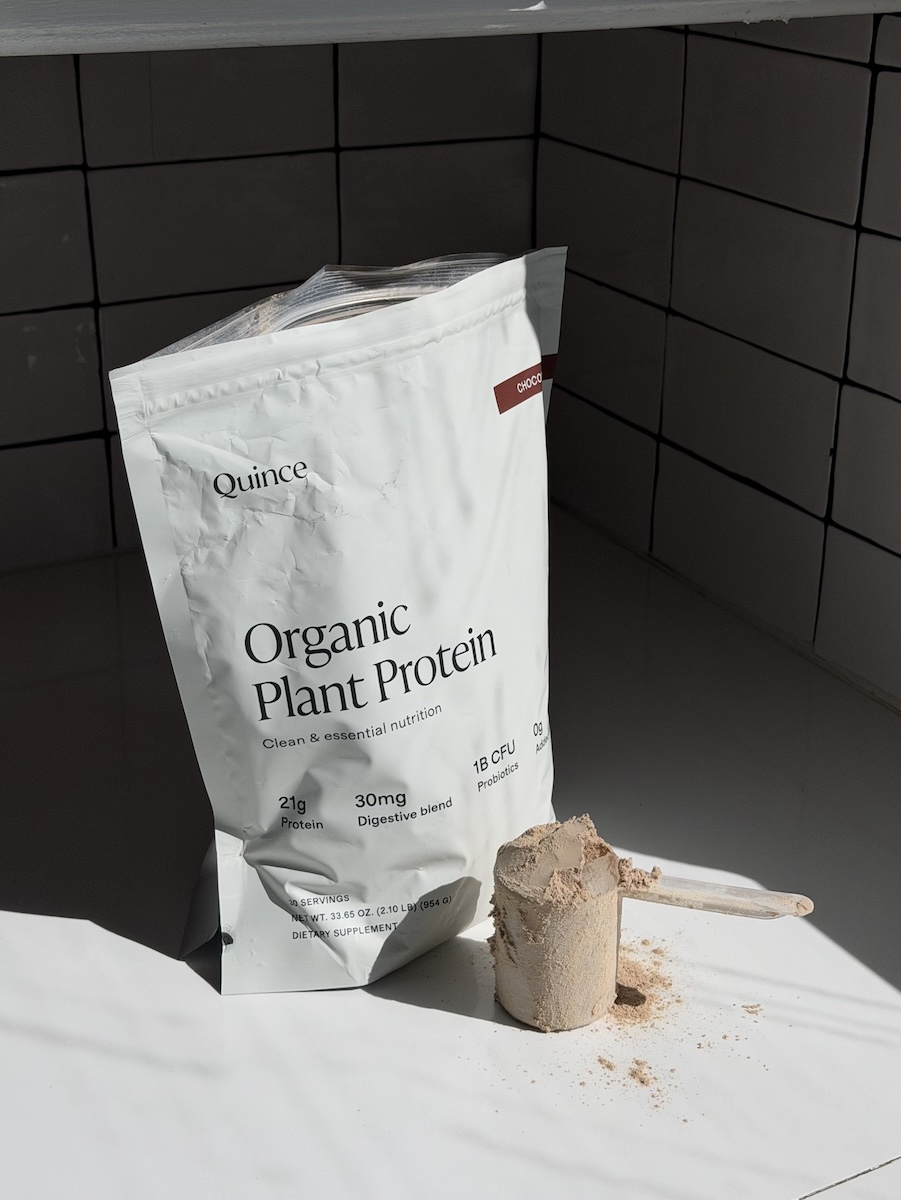 A bag of Quince Organic Plant Protein powder stands on a white tiled counter with a full scoop of powder and some powder spilled beside it.
