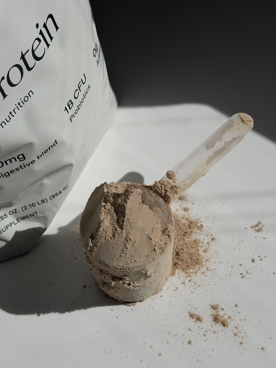 A scoop of brown protein powder sits on a white surface next to a partially visible protein powder bag.