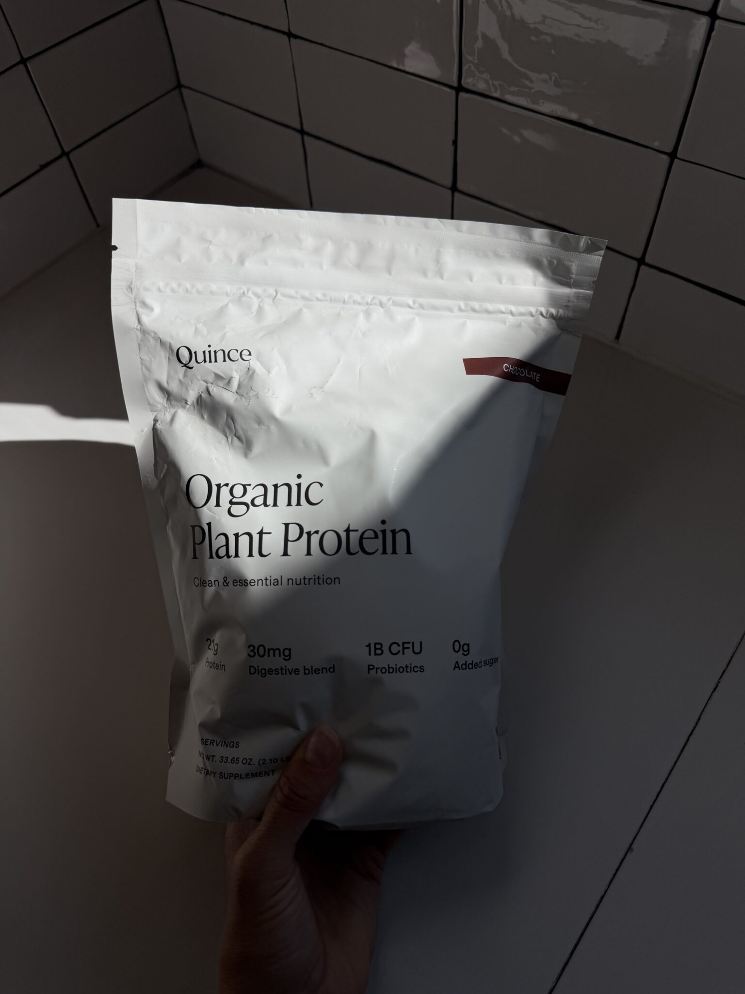 A hand holds a bag of Quince Organic Plant Protein against a white tiled wall and surface. The packaging lists features like digestive blend, probiotics, and no added sugar.