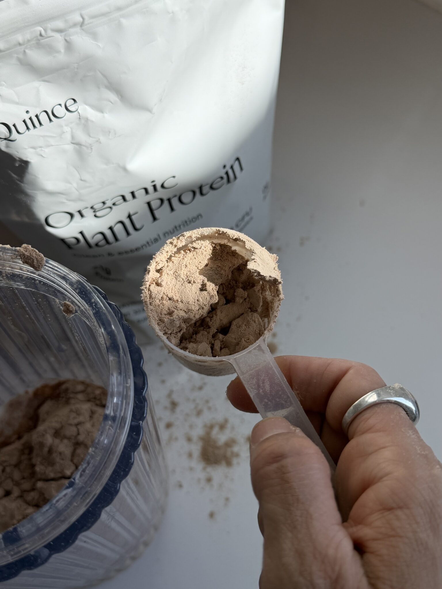 A hand holding a scoop of brown plant protein powder above a container, with a bag labeled "Organic Plant Protein" in the background.