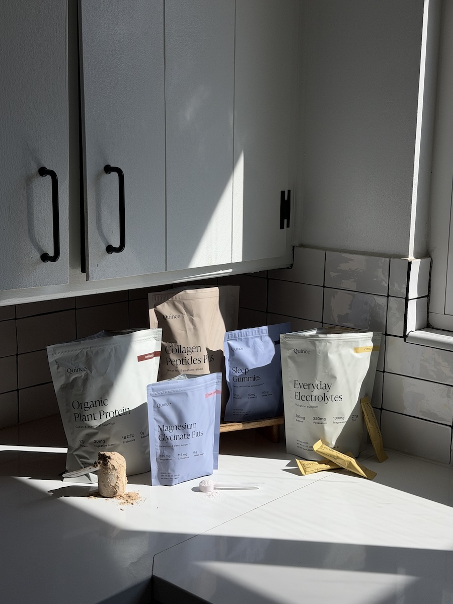 Five health supplement packages are displayed on a white kitchen counter under natural light, with two scoops of powder and three stick packs in front.