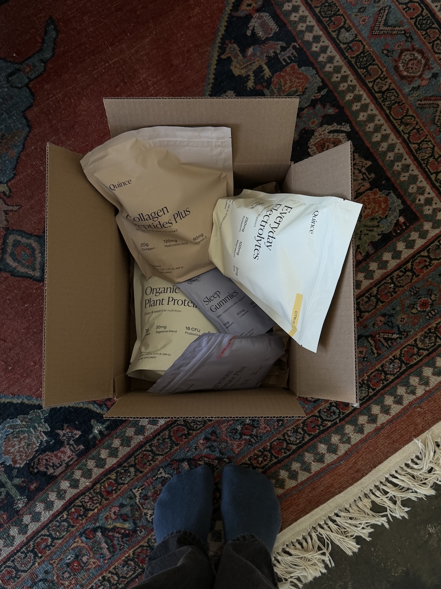Open cardboard box containing several large packets of protein and supplements sits on a patterned rug, with a person's socked feet visible at the bottom of the image.