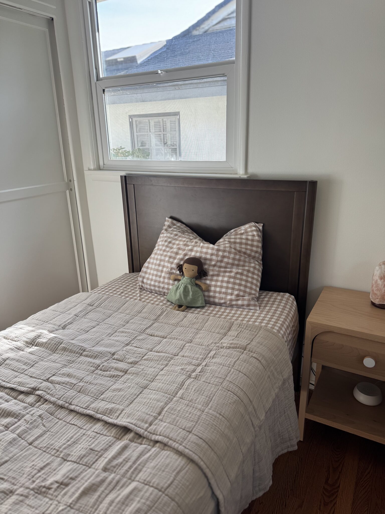 A neatly made single bed with a checkered pillow and a doll on top, next to a wooden nightstand, in a bright room with a window.