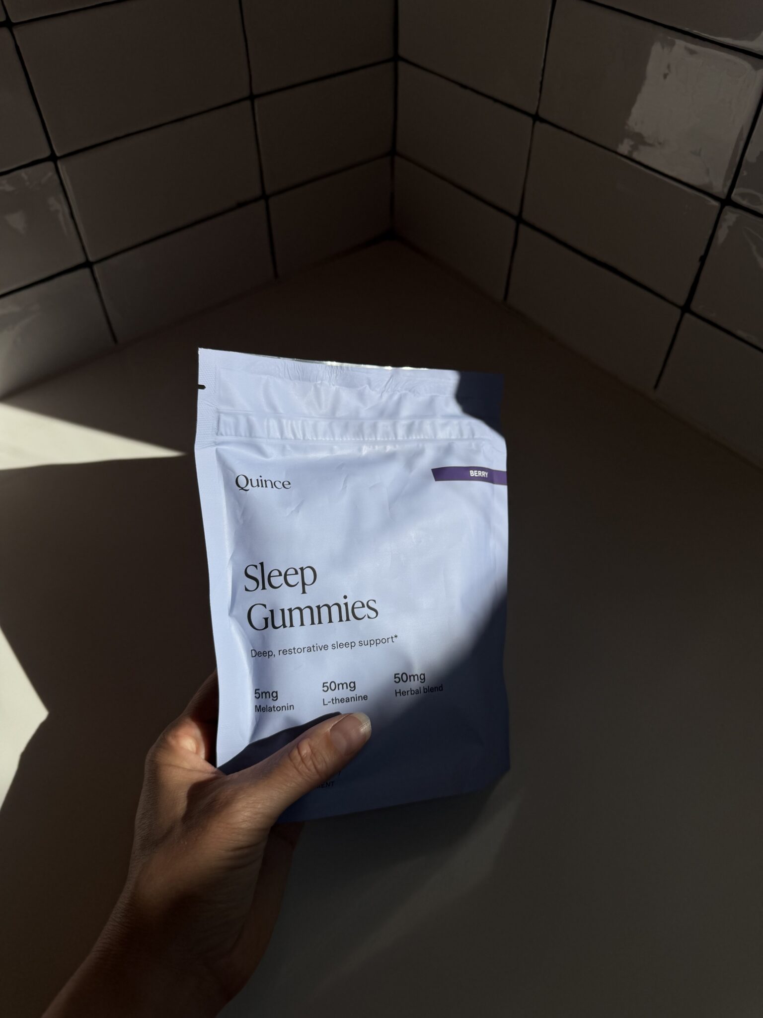 A hand holds a packet of Quince Sleep Gummies in a sunlit room with white tiled walls and a white countertop.