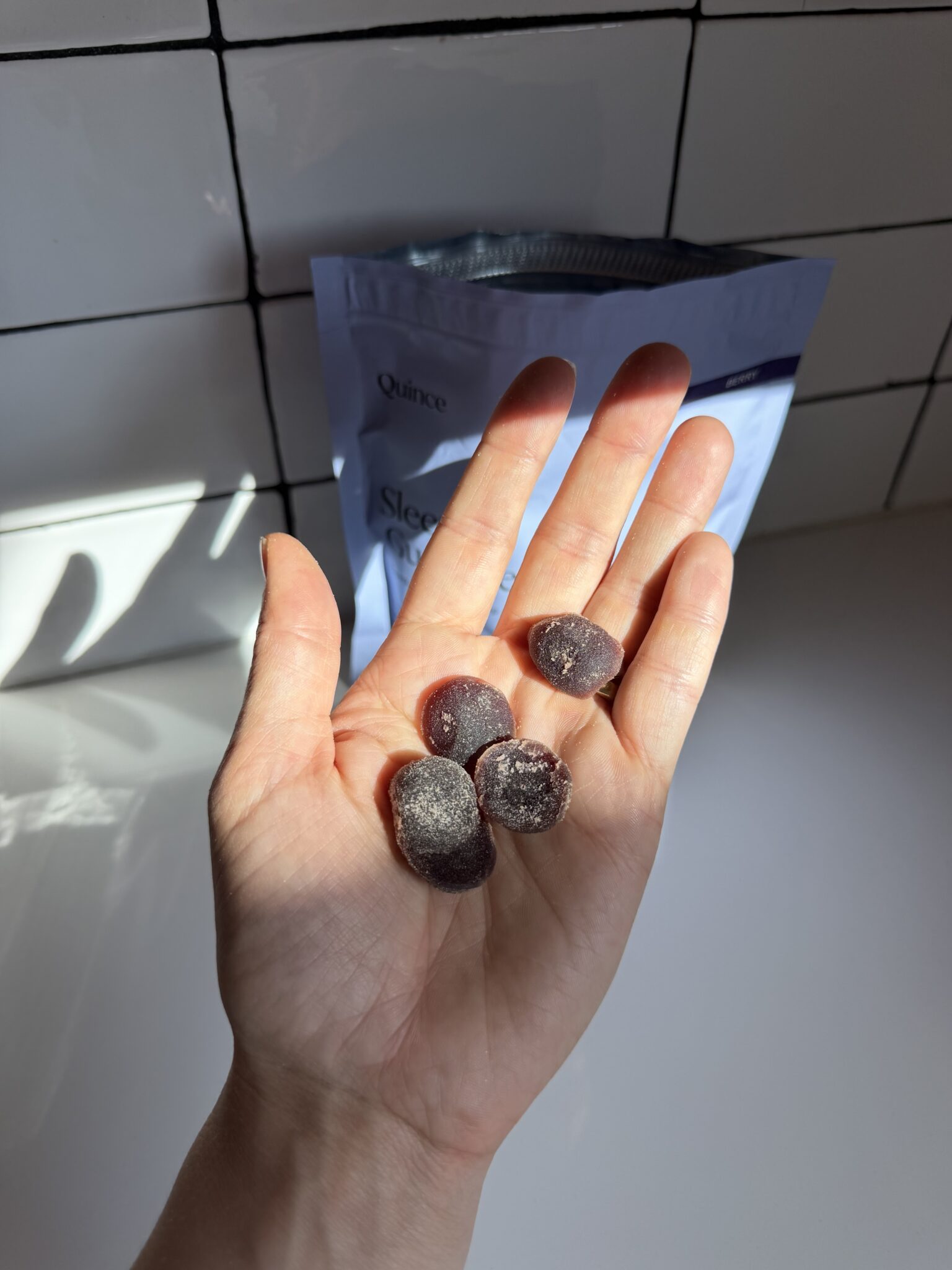 A hand holds five dark, round gummies with a powdery coating; a partially visible blue resealable bag is in the background on a white tiled surface.
