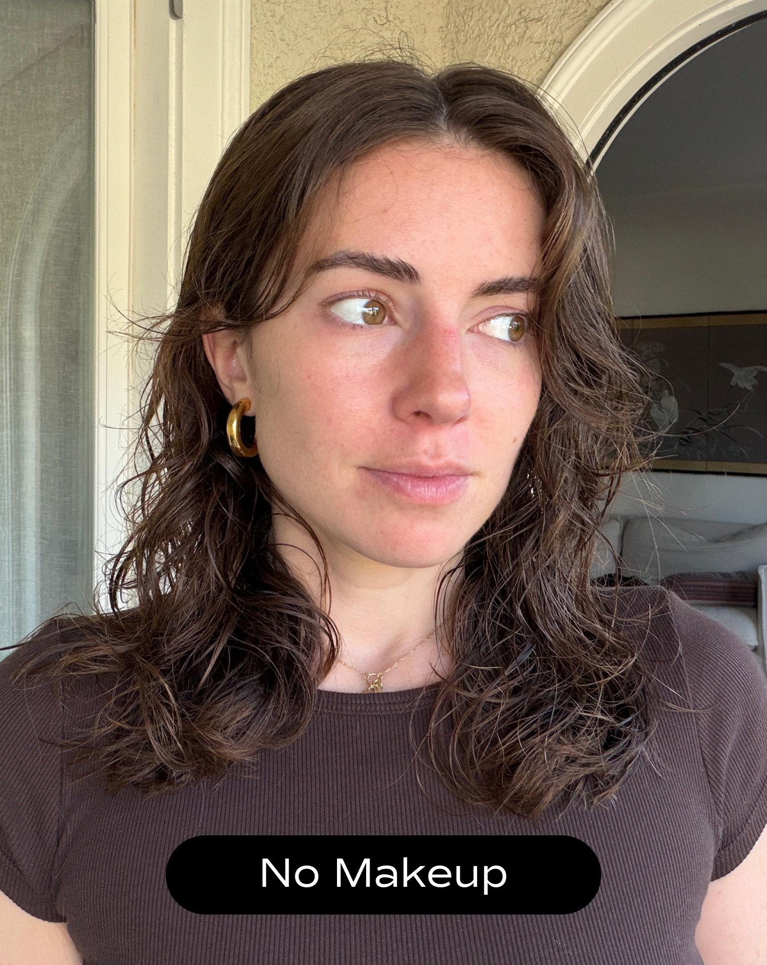 A woman with wavy brown hair, wearing gold hoop earrings and a brown shirt, stands indoors with a neutral expression. The text "No Makeup" appears at the bottom.