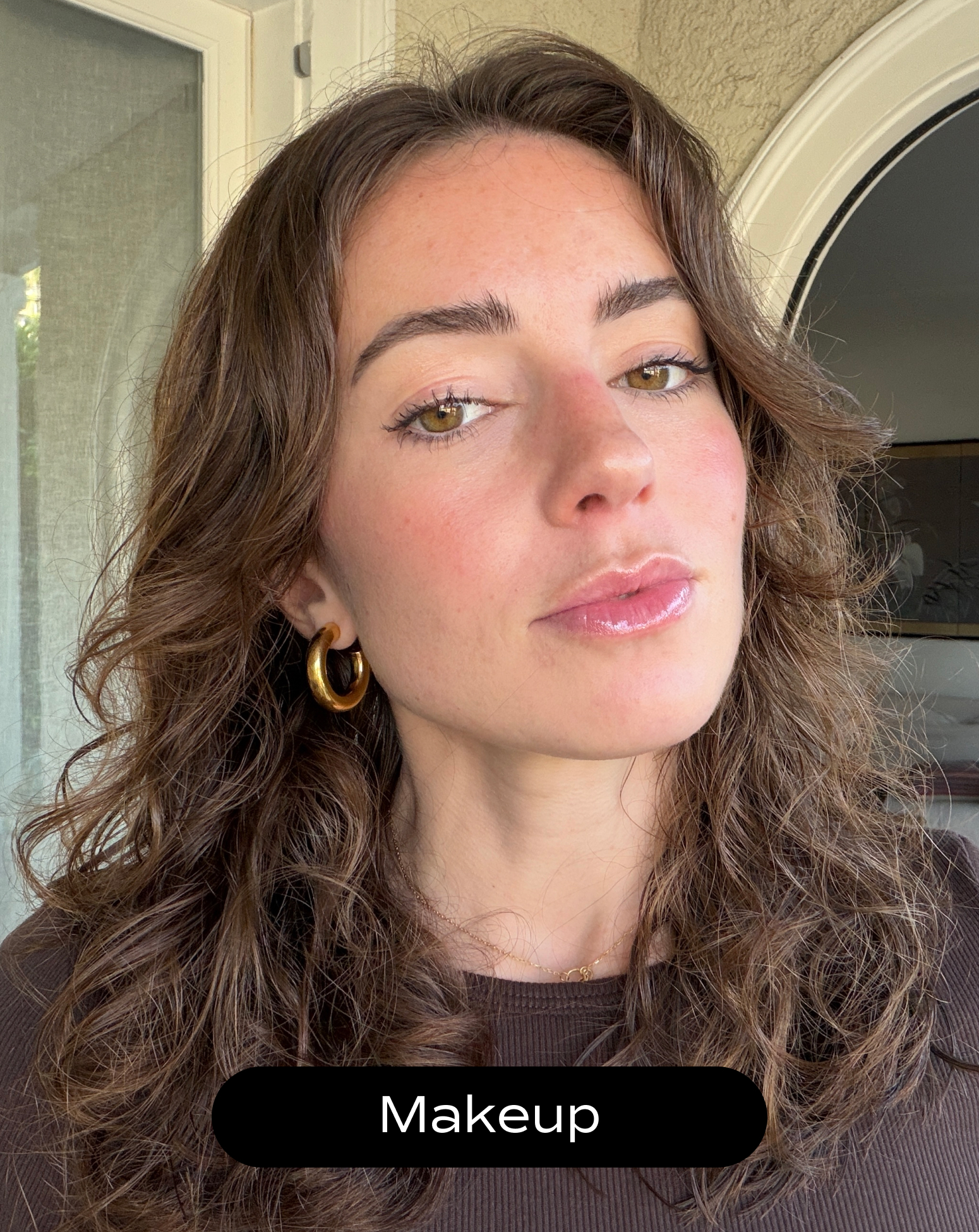 A woman with wavy brown hair wearing gold hoop earrings, natural makeup, and a brown top stands indoors near a door. The word "Makeup" appears at the bottom of the image.