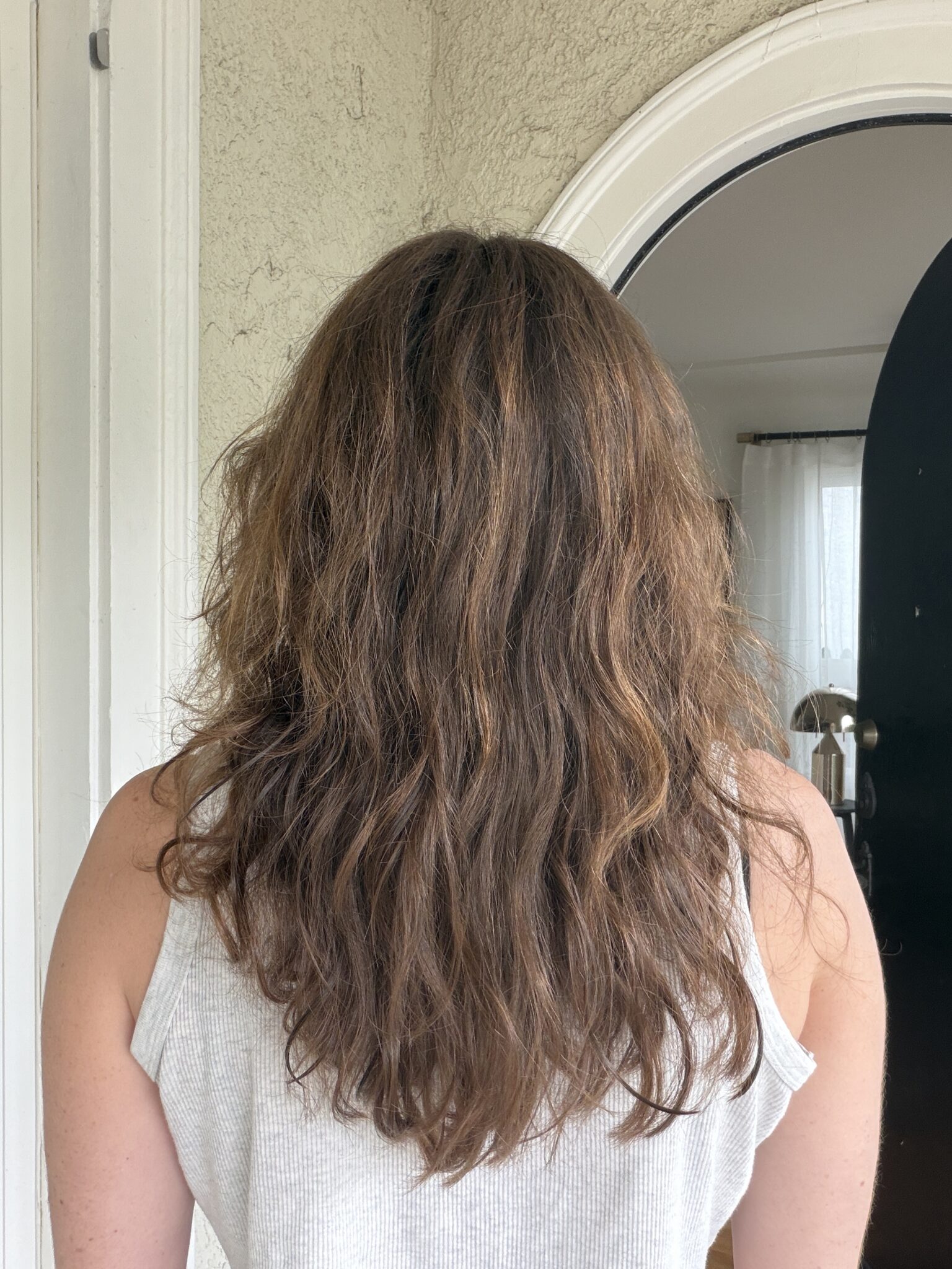 A person with shoulder-length, wavy brown hair is standing indoors facing away from the camera, wearing a light-colored sleeveless top.