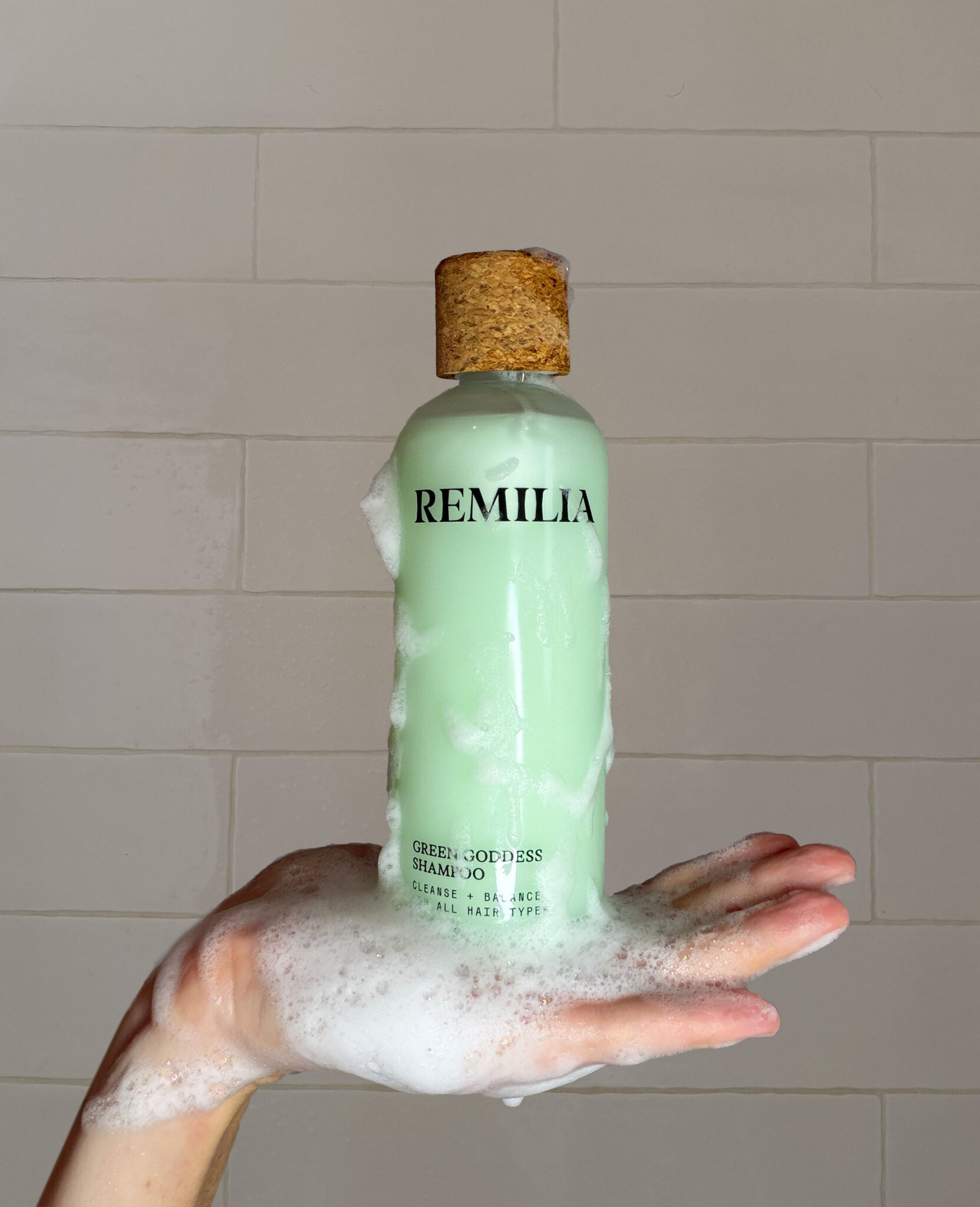 A hand covered in soap suds holds a green bottle of REMILIA Green Goddess Shampoo against a tiled bathroom wall.
