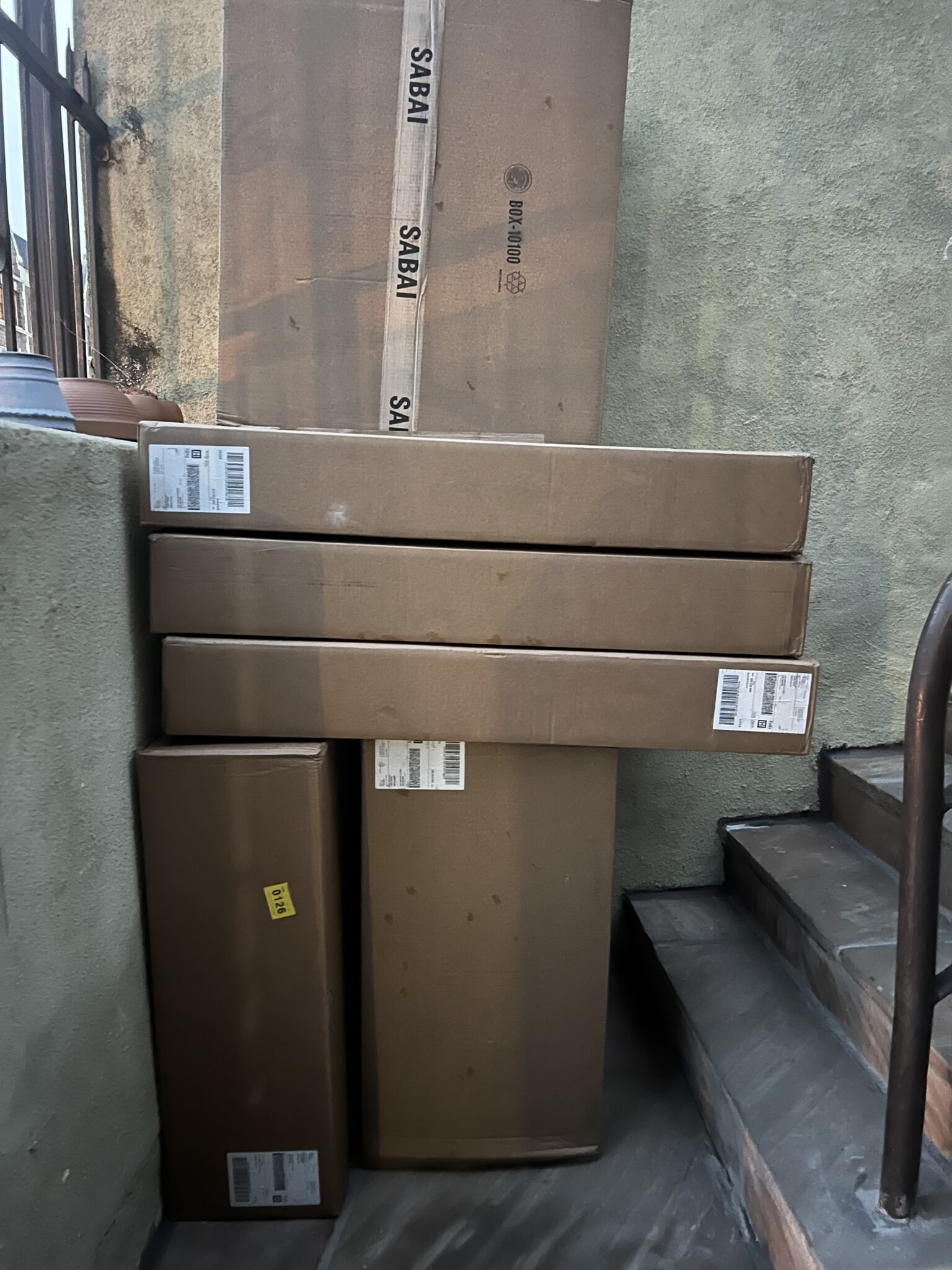 Five cardboard shipping boxes of various sizes stacked next to a staircase and green wall, with shipping labels and a "SABA" logo visible on some boxes.