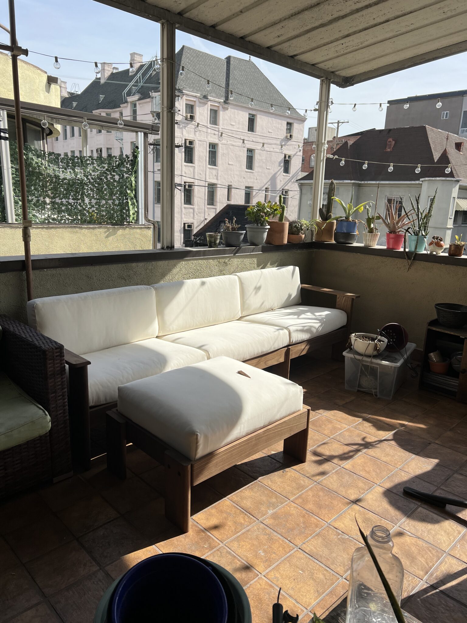 A sunlit balcony with a white cushioned outdoor sofa, potted plants on the ledge, string lights, and tiled floor overlooking buildings.