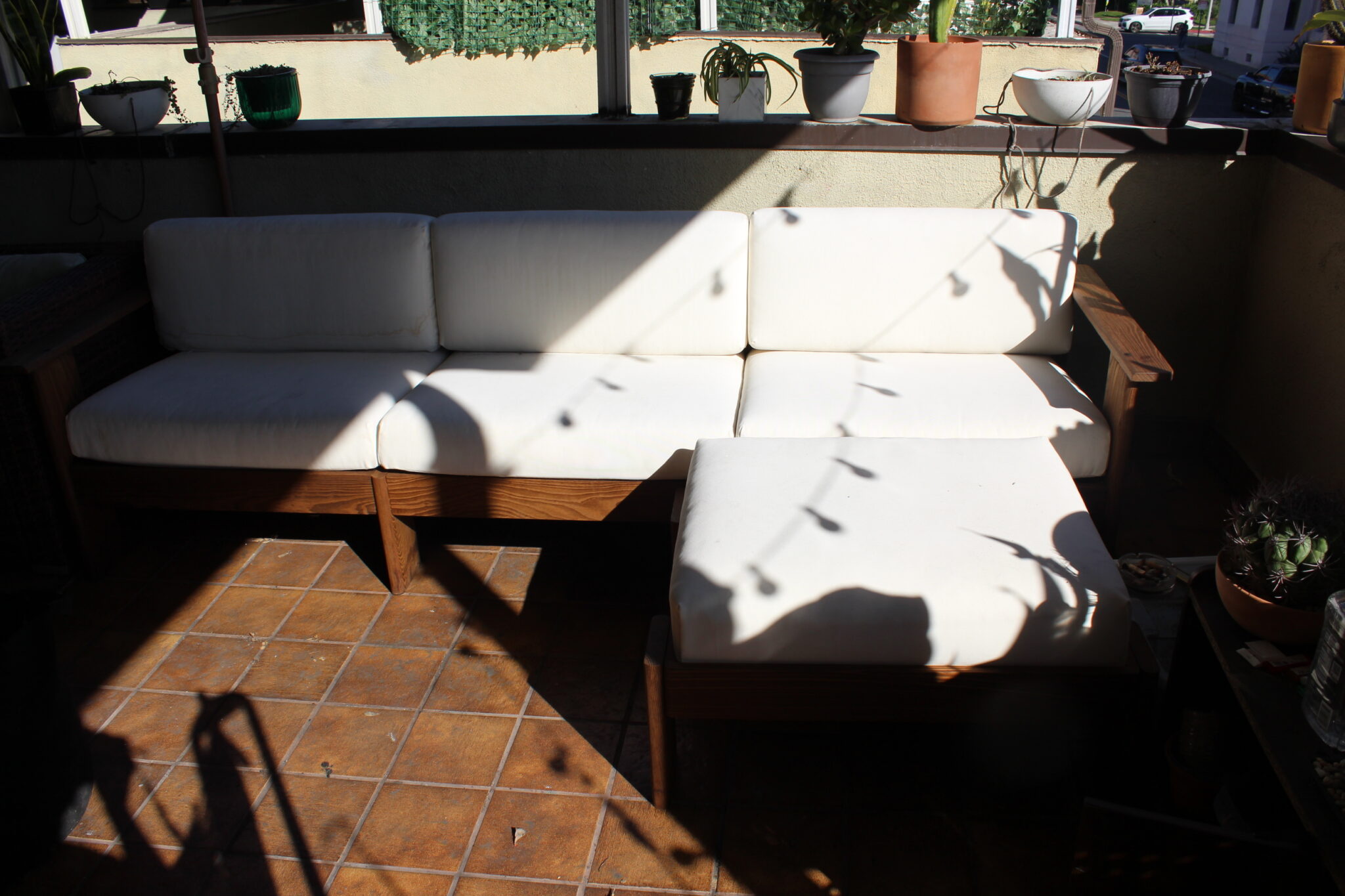 Wooden outdoor sofa with white cushions and an ottoman on a tiled patio, surrounded by potted plants, partially shaded by sunlight and shadows.