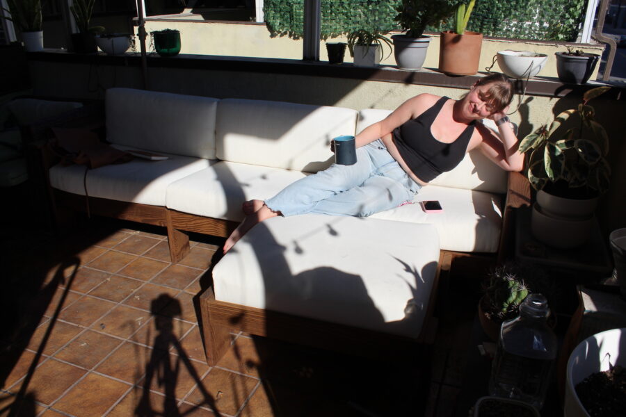 A woman lounges on a white outdoor sofa, holding a blue cup. There are plants around her and strong sunlight casts shadows on the tiled floor and furniture.
