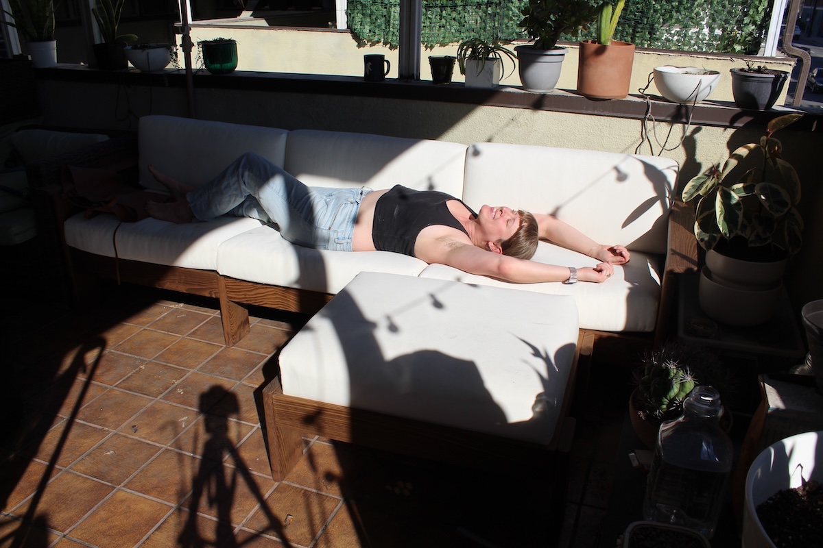 A woman is lying on a white sofa with her arms stretched out, surrounded by indoor plants and sunlight streaming through a window.